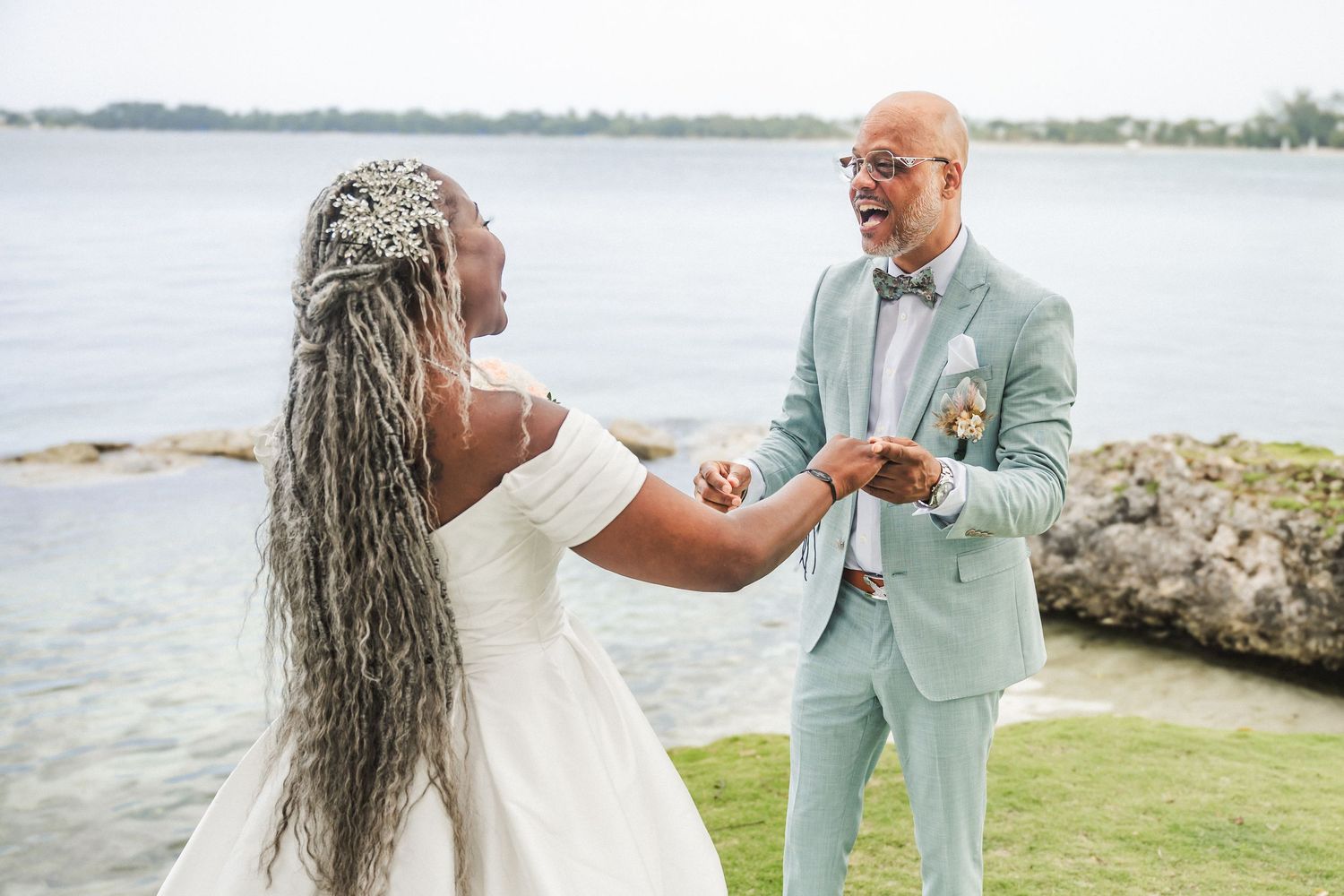 A happy couple in a mint suit and white wedding dress dance and laugh together by a waterfront on their wedding day.