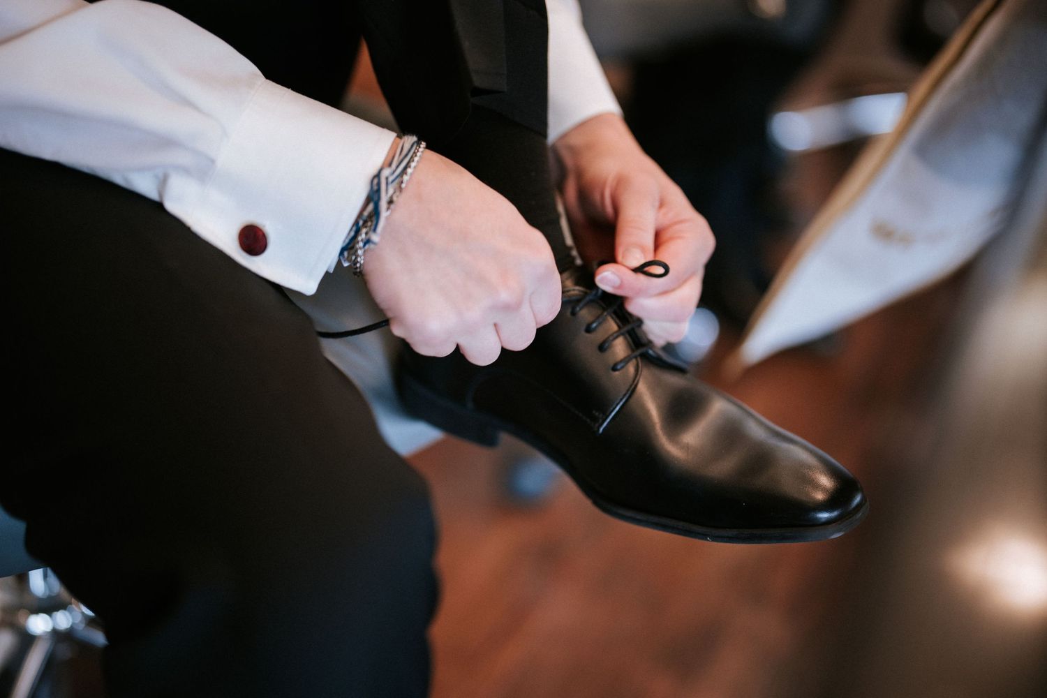 Person tying laces on polished brown leather dress shoes while wearing formal white shirt with cufflinks.