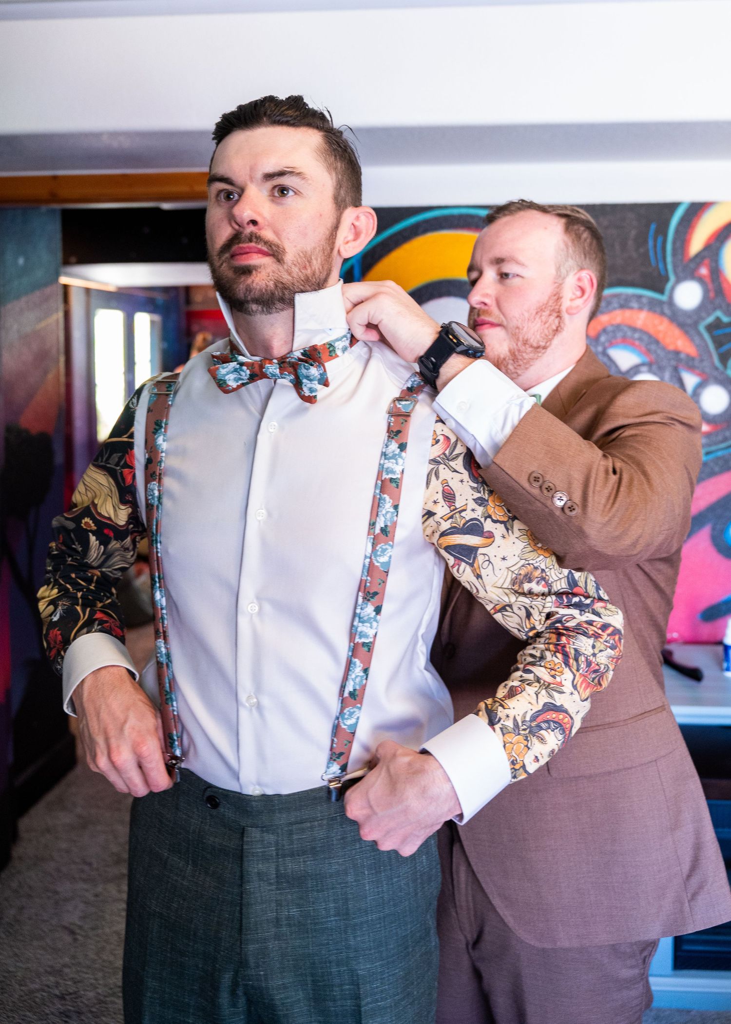 Two people in formal attire help with adjusting colorful floral suspenders against a vibrant graffiti wall background.