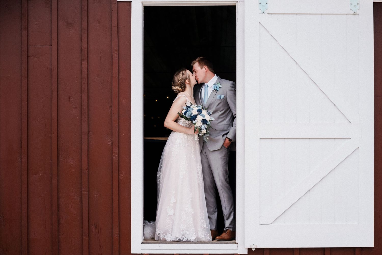 A couple in wedding attire share an intimate moment in the doorway of a rustic red barn.
