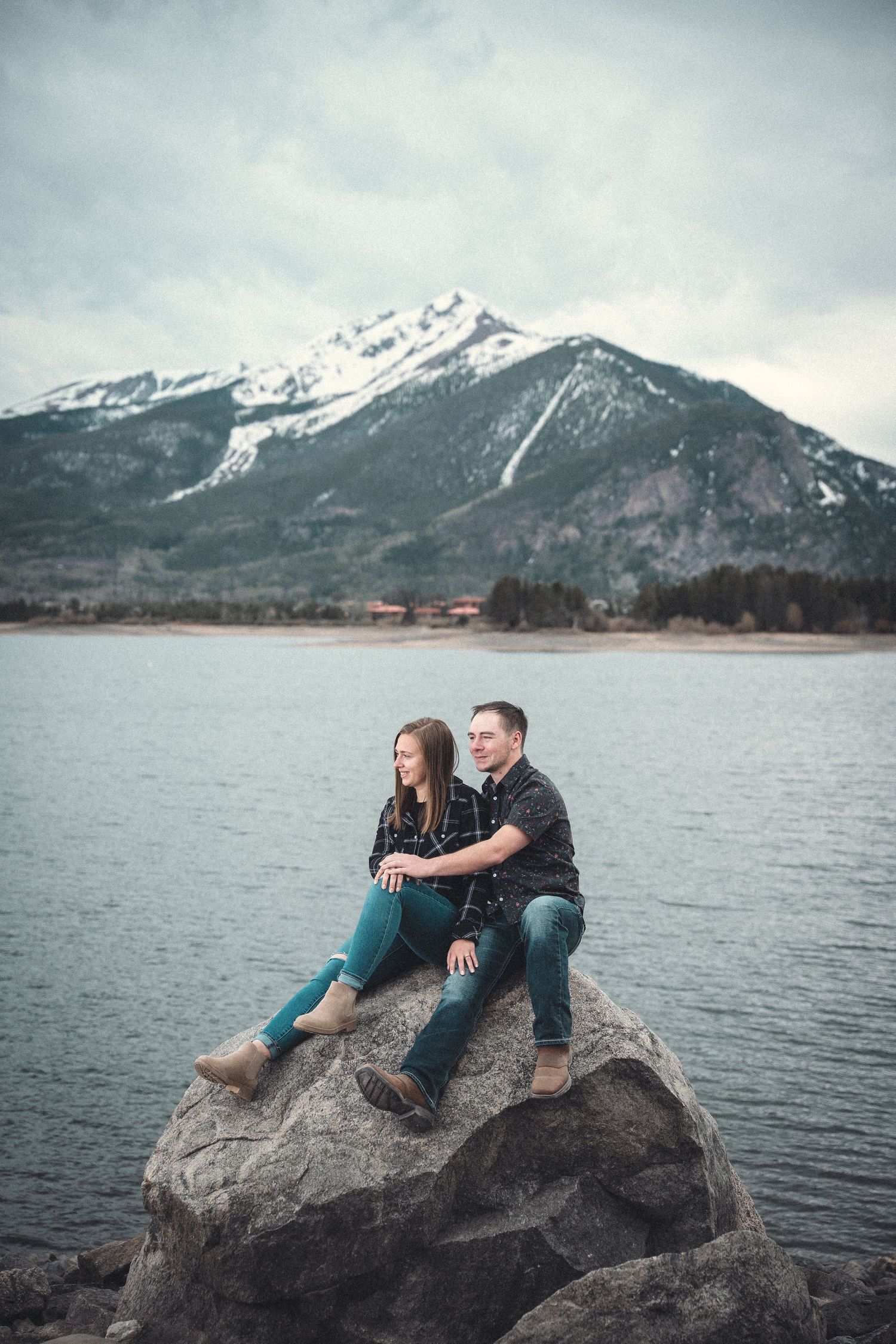 Two people sit together on a large rock by a lake with snow-capped mountains in the background.
