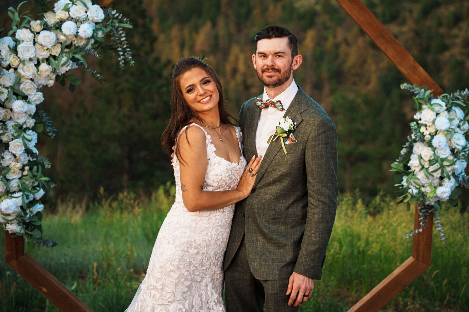 A couple poses under a wooden hexagonal arch decorated with blue and white flowers at an outdoor wedding ceremony.