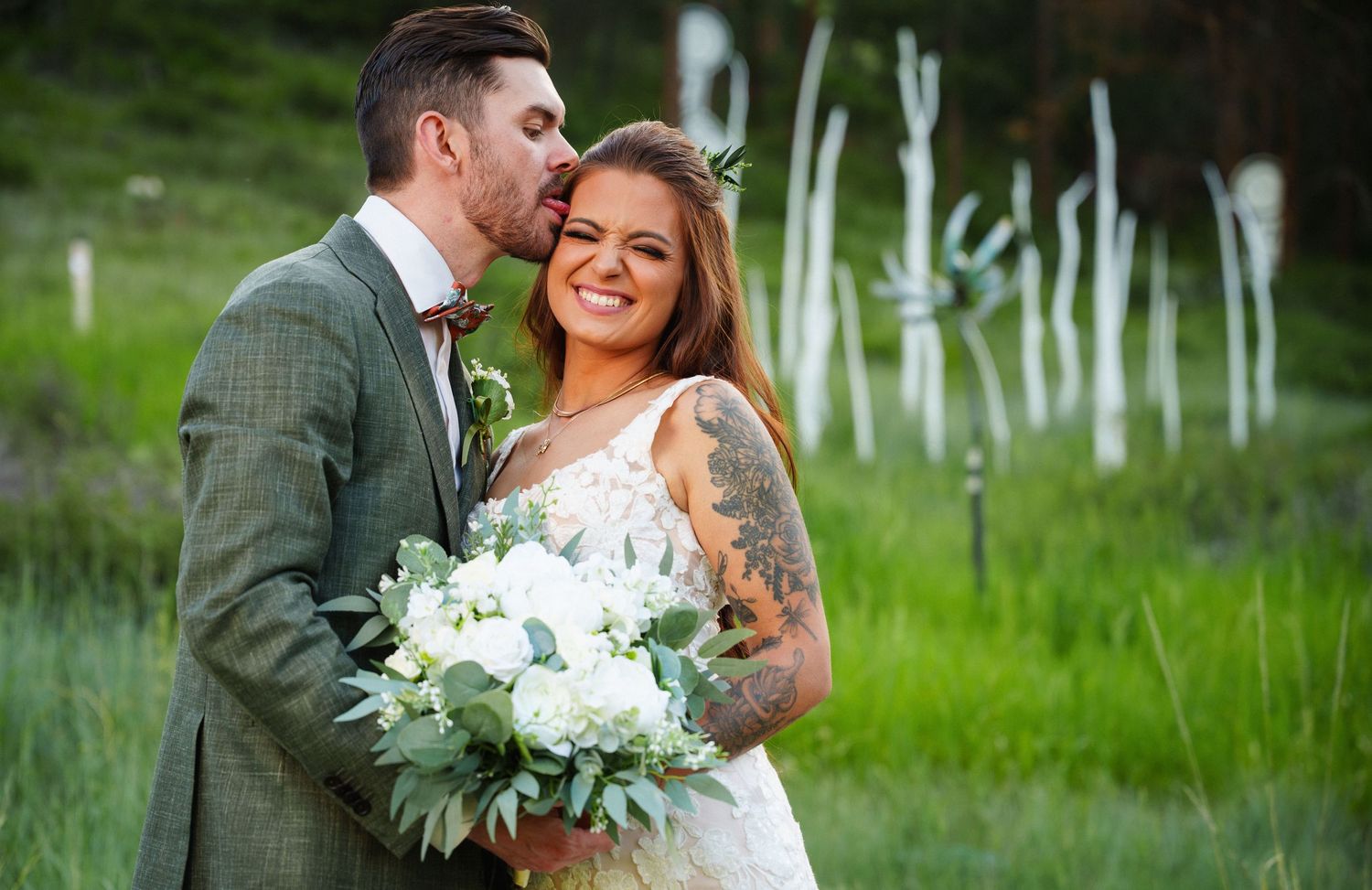 A couple in wedding attire share a romantic moment in a green meadow with white floral bouquet.