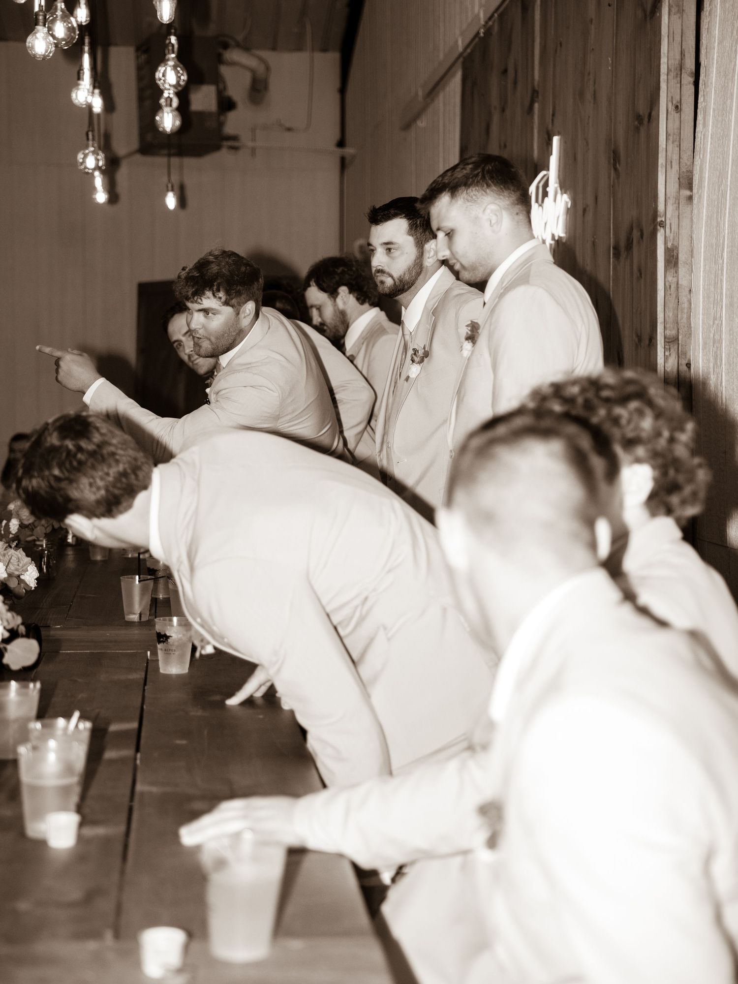 A vintage sepia-toned panoramic photo shows groomsmen wearing white suits leaning diagonally at a wedding reception.