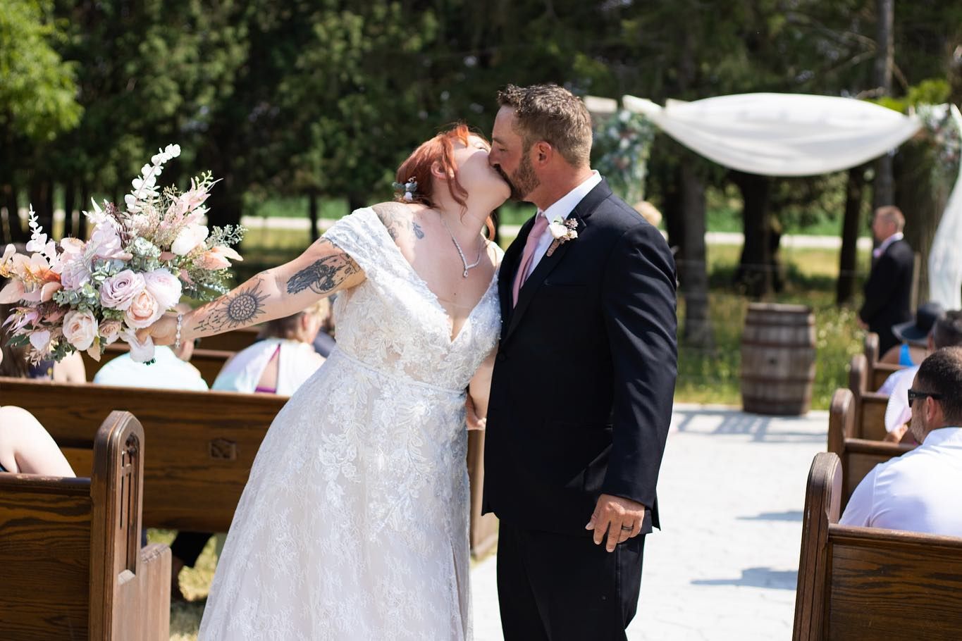 A wedding couple shares an intimate kiss in front of rustic wooden benches decorated with floral arrangements.