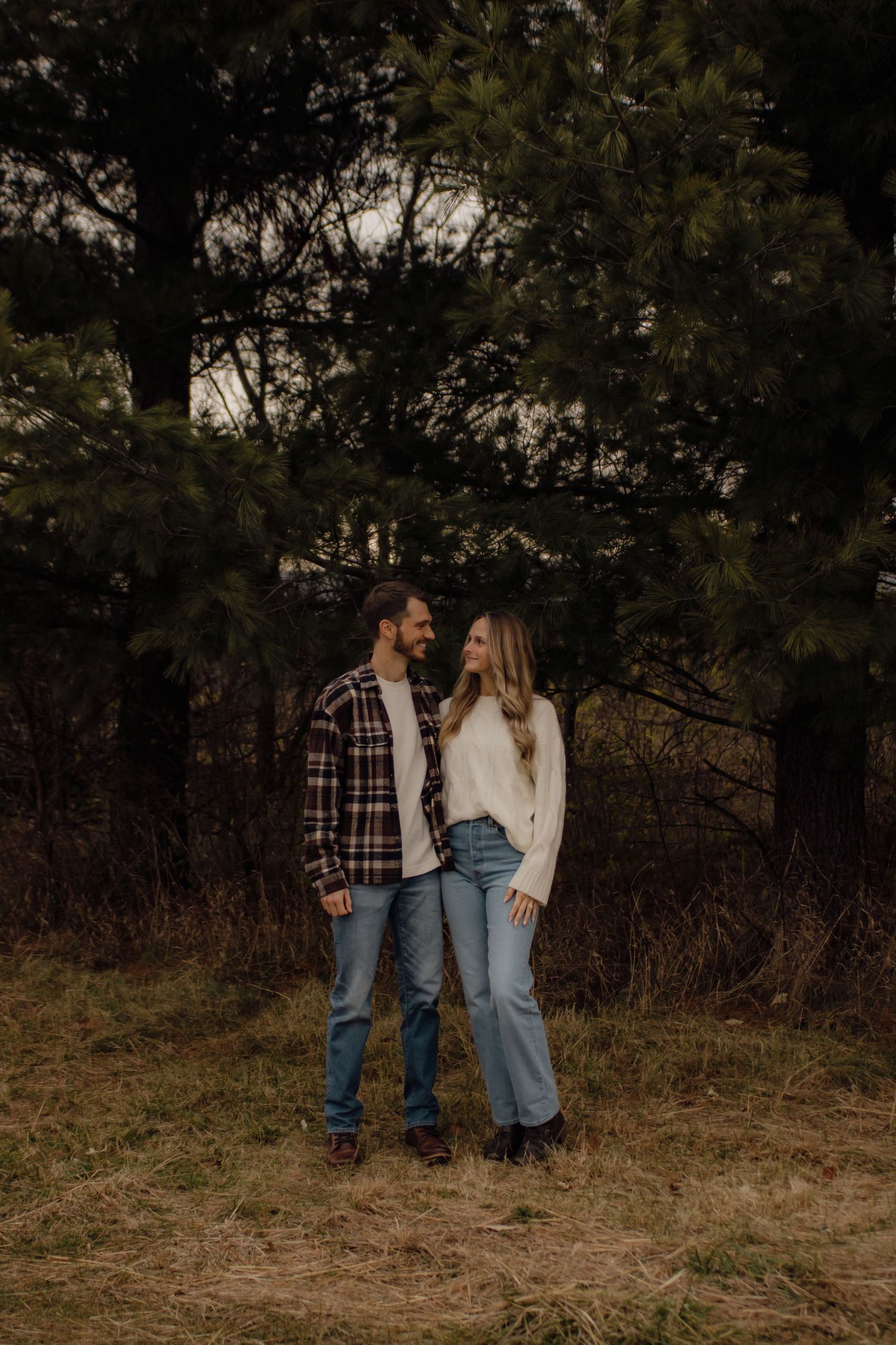 A casual outdoor couple portrait with plaid shirt and cream sweater against autumn foliage.