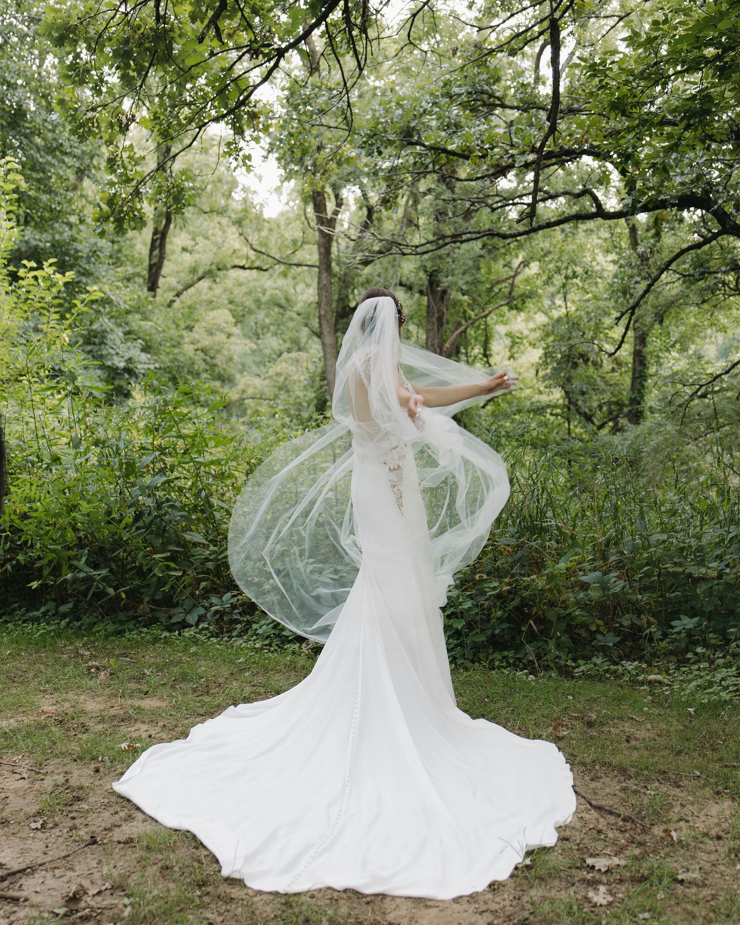 A flowing white wedding gown and veil catch the breeze in a lush green garden setting.