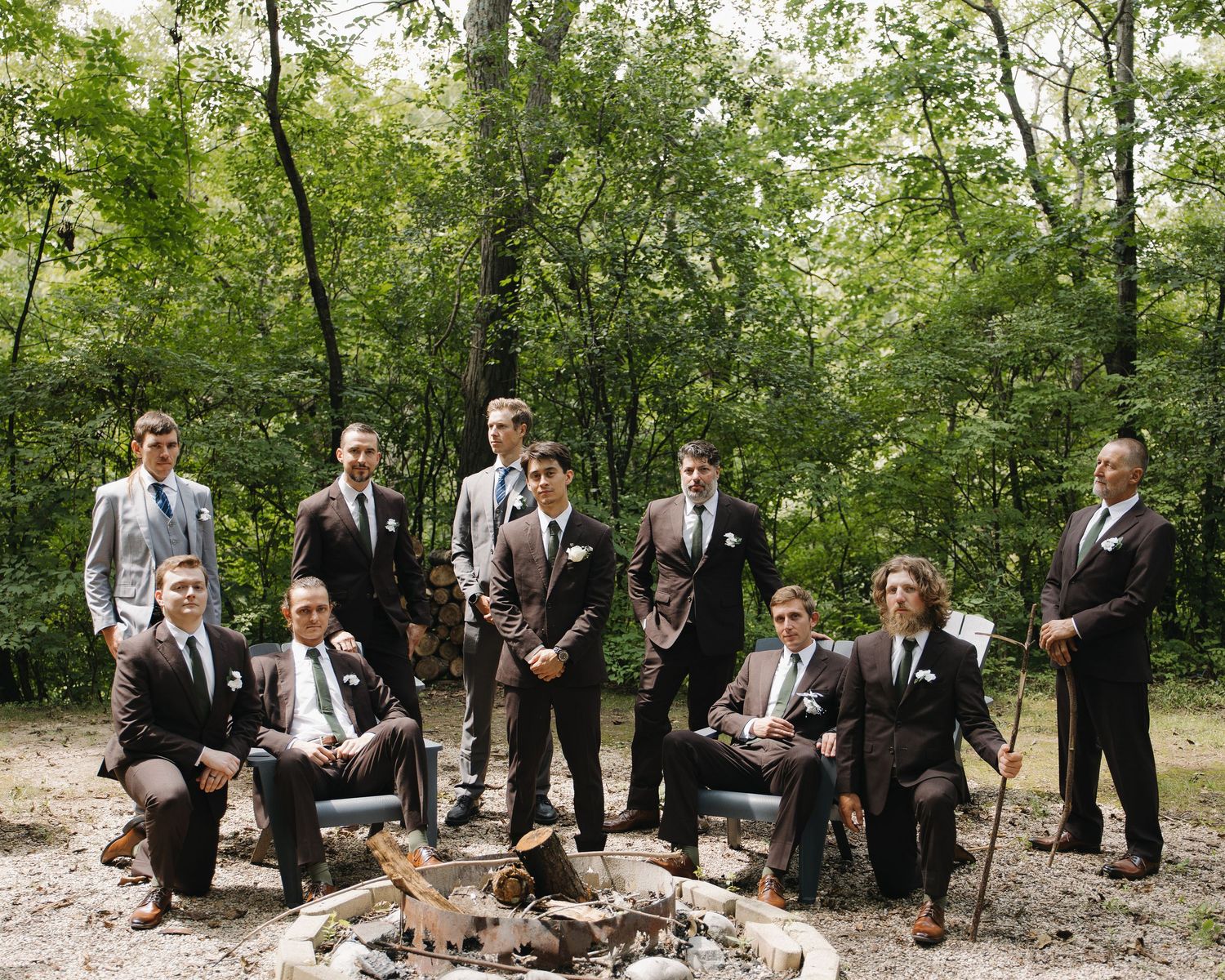 A wedding party in grey and black suits gather around a rustic outdoor firepit in a wooded setting.