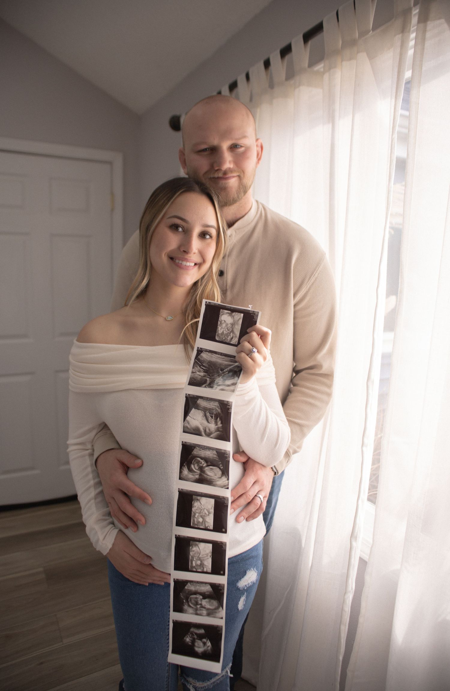 Couple displays strip of ultrasound photos while standing by window.