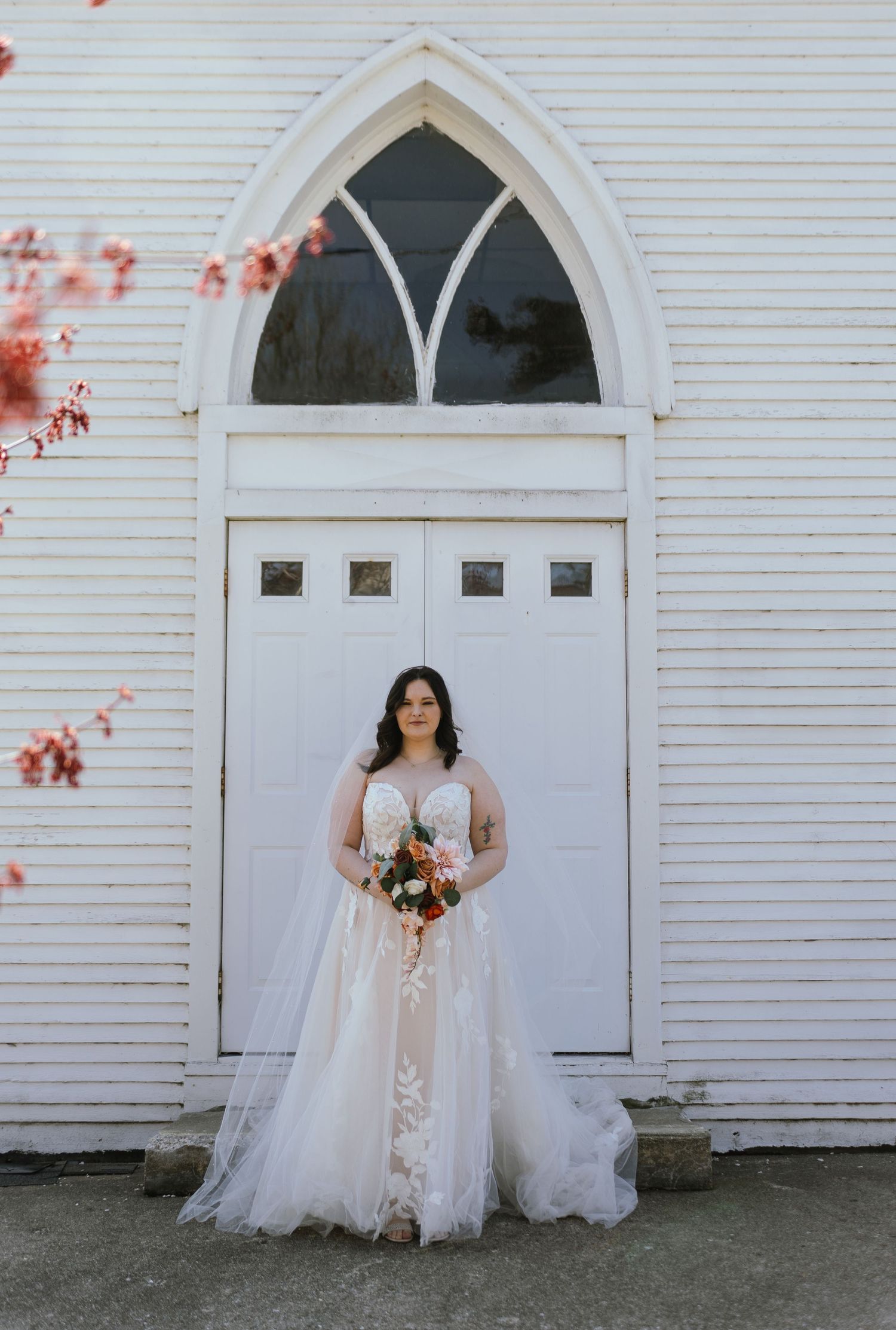 A bride in a lace gown holding a bouquet in front of a white church with gothic windows.