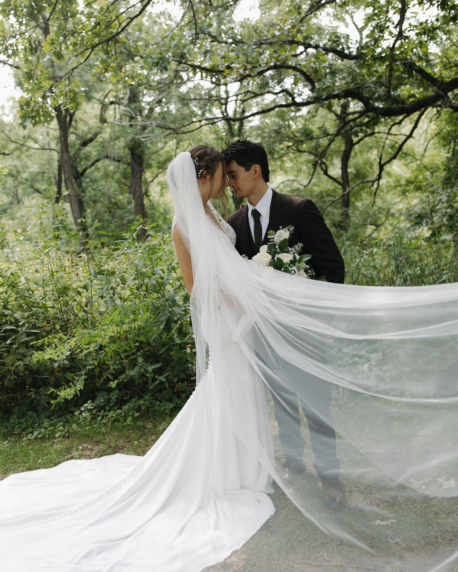 Newlywed couple shares romantic moment in nature setting with flowing white wedding dress and long fluttering veil.