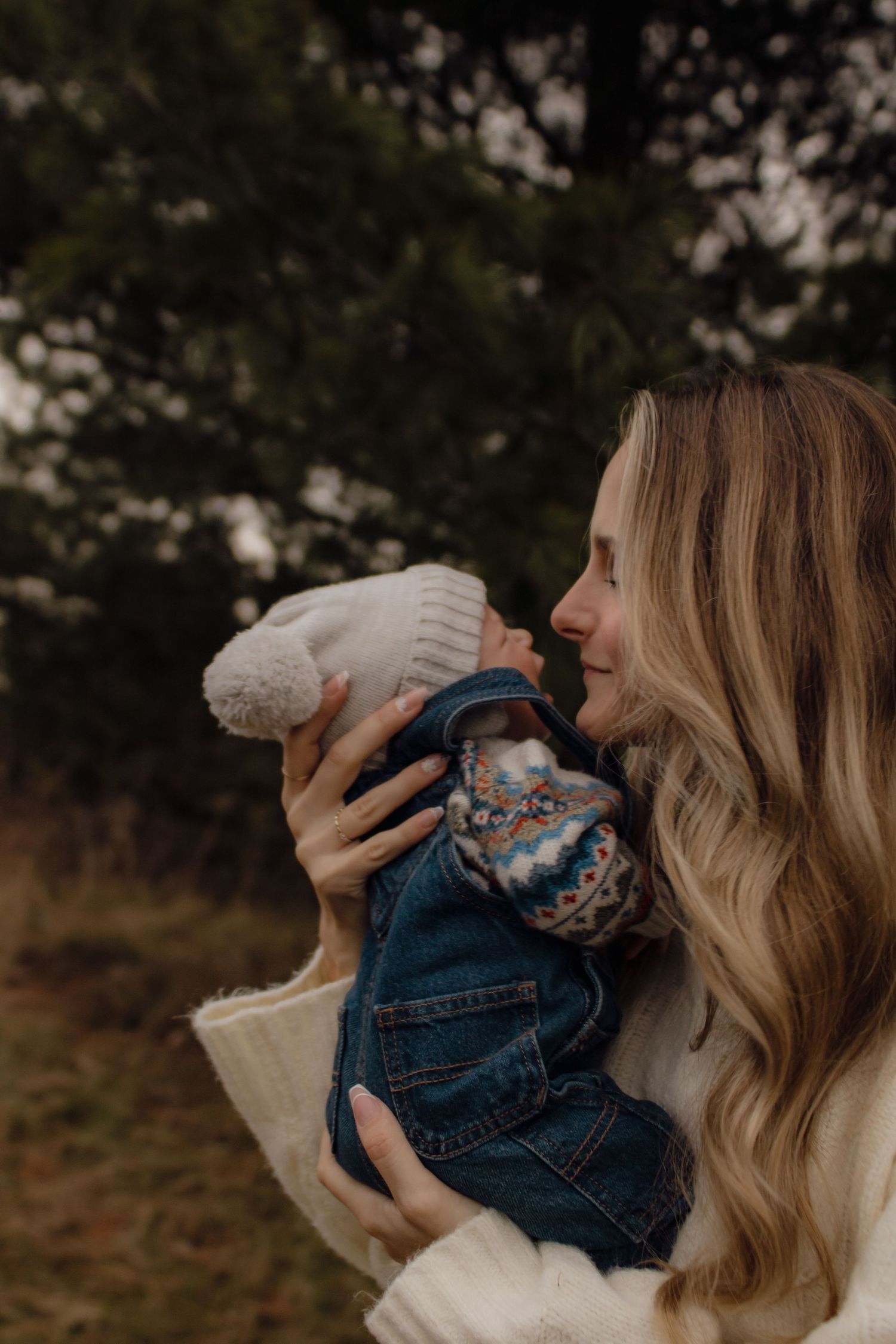 Woman in white sweater holding small infant in denim outfit outdoors on autumn day.