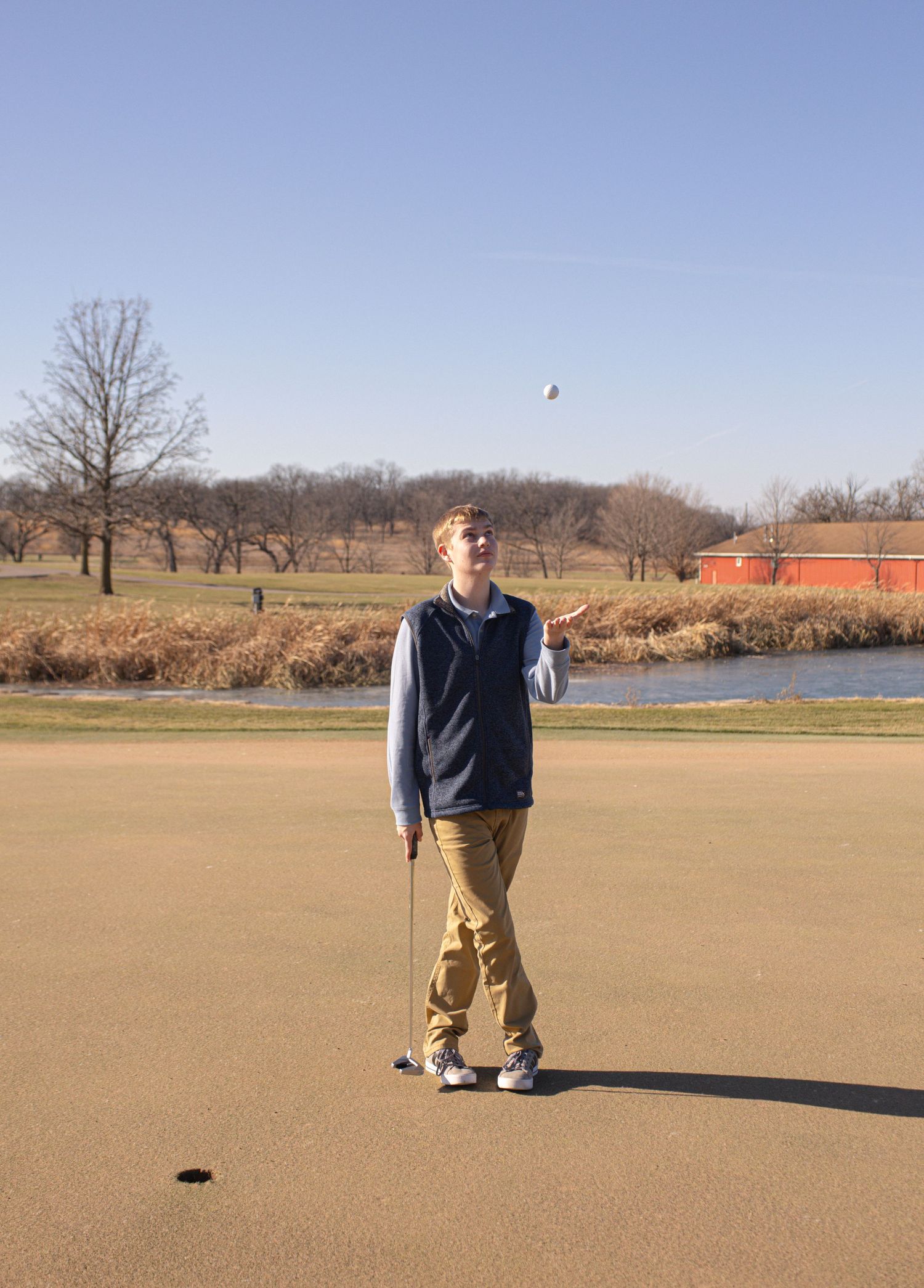 A person stands on a putting green with a golf ball, surrounded by autumn scenery and a pond.