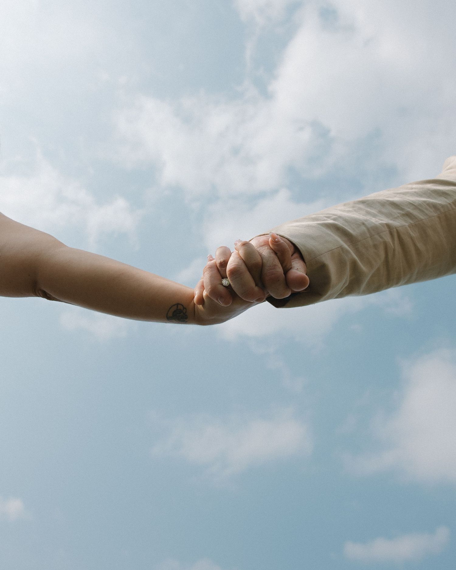 Two hands reaching out to hold each other against a soft cloudy blue sky.