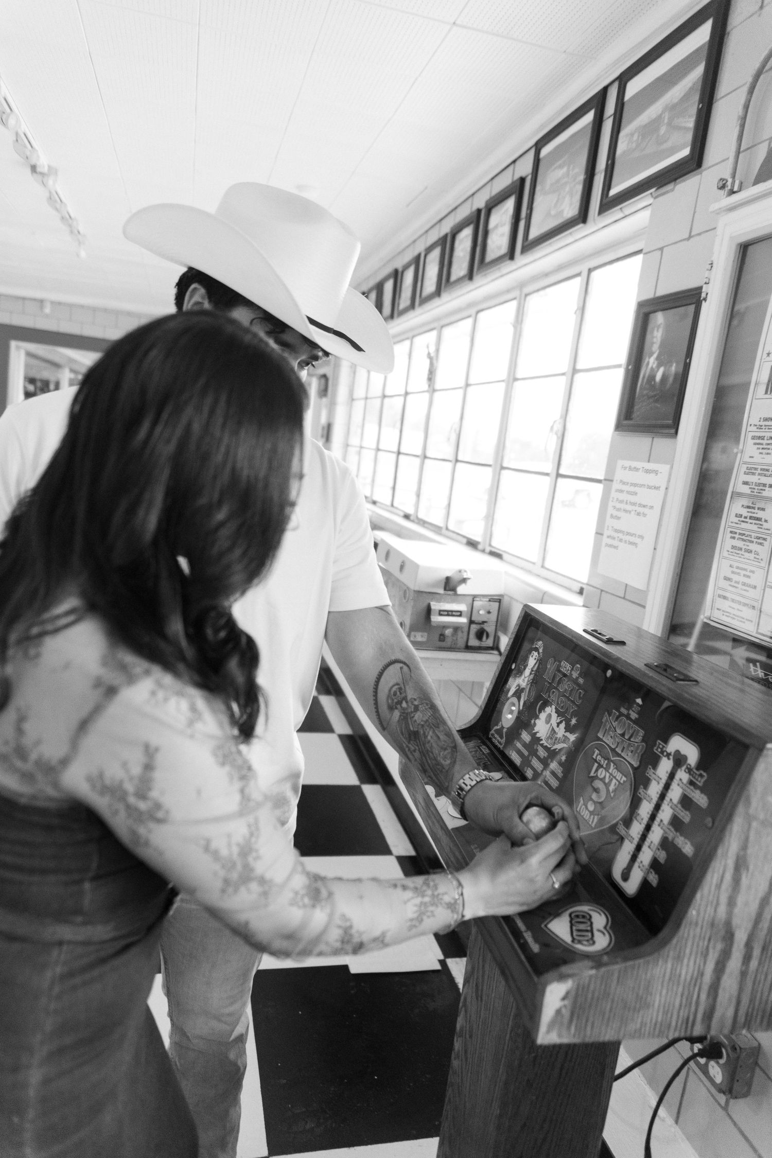 Person in cowboy hat and jeans examines an arcade game control panel in black and white photograph.