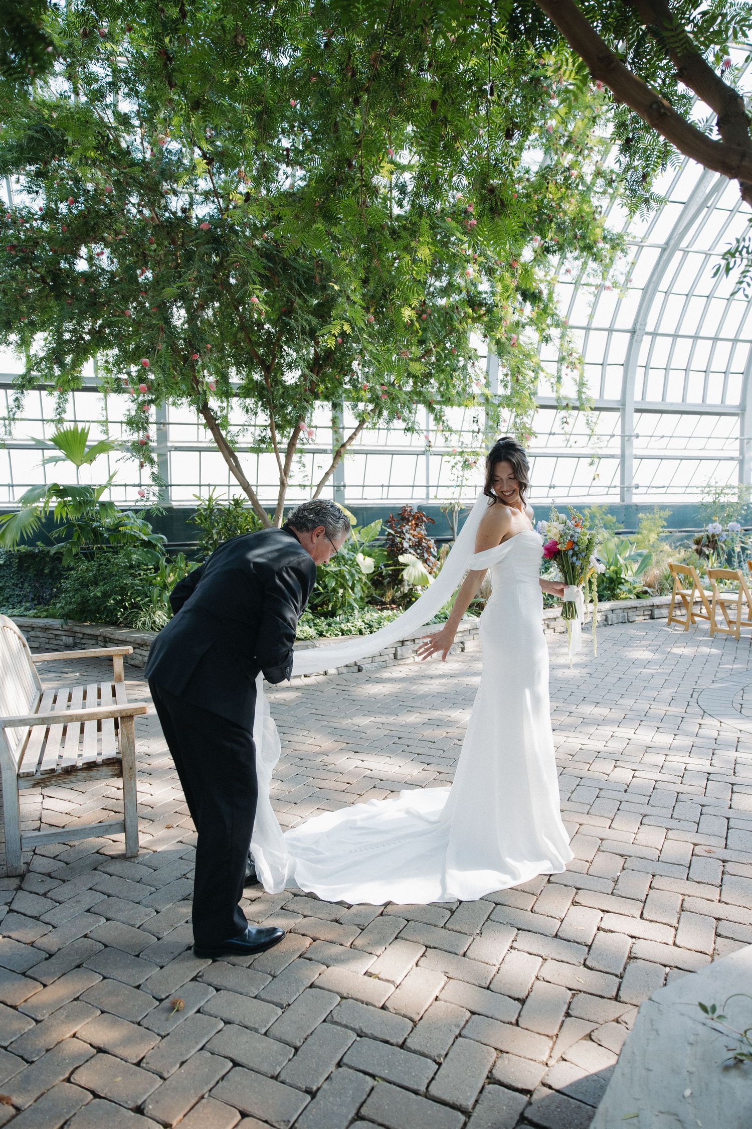 A wedding couple shares a tender moment in a sunlit greenhouse conservatory with brick pavers and lush greenery overhead.