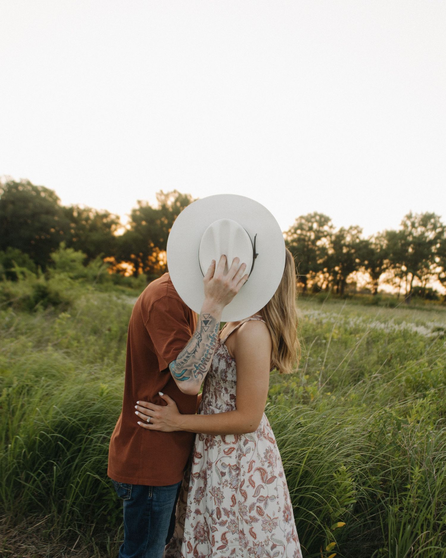 A couple stands close together in a grassy field at sunset wearing brown and white floral clothing.
