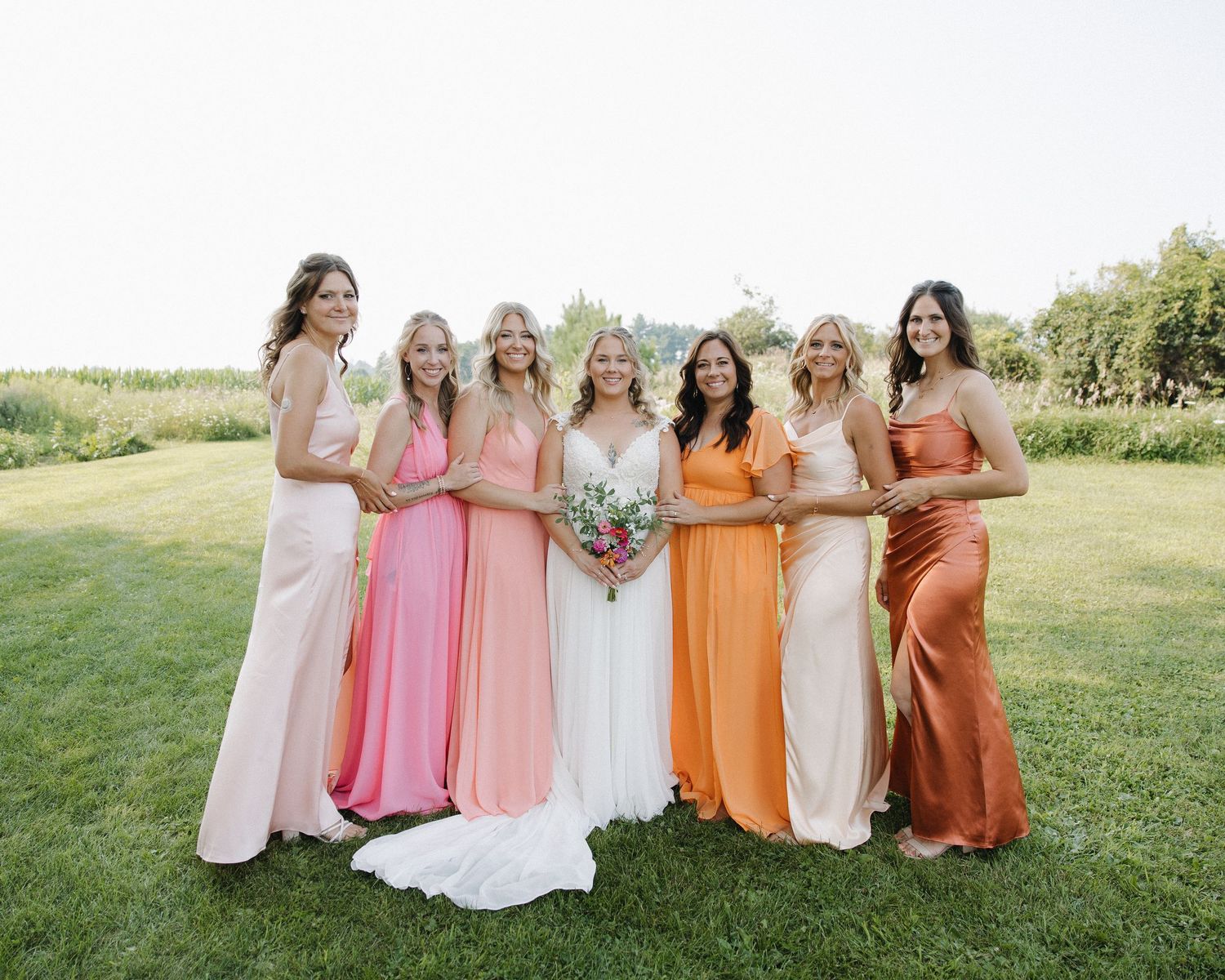 Wedding party in colorful pastel and earth tone dresses pose together on a grassy hill during a summer celebration.