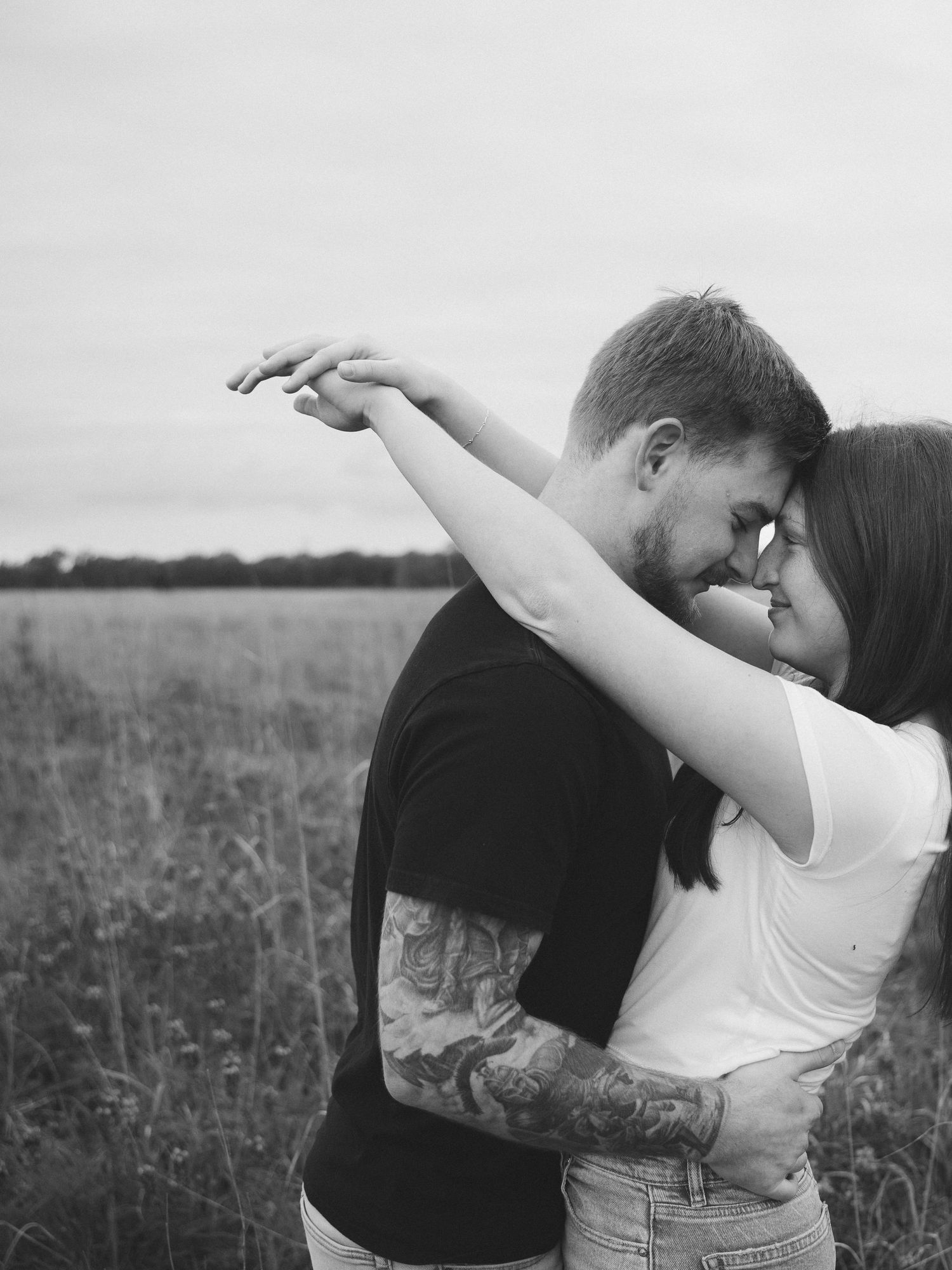 A romantic black and white portrait of a couple embracing in a field, with the woman's arm raised gracefully.