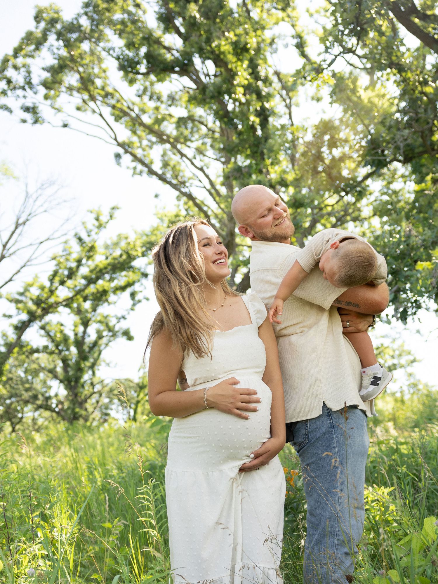 A joyful family plays together outdoors in a sunlit meadow with green trees and tall grass on a summer day.