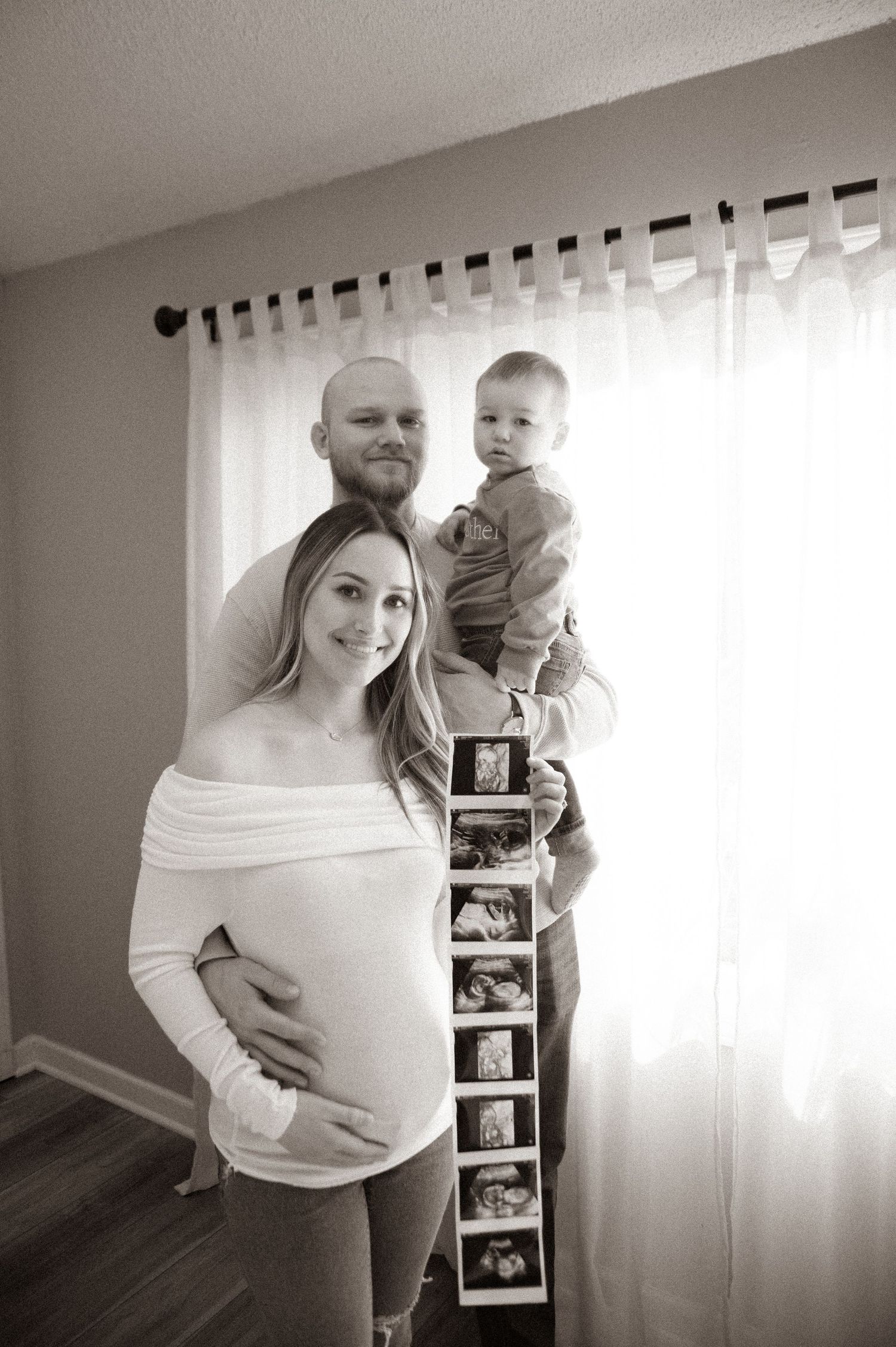 Family poses with ultrasound photo strip in front of window.
