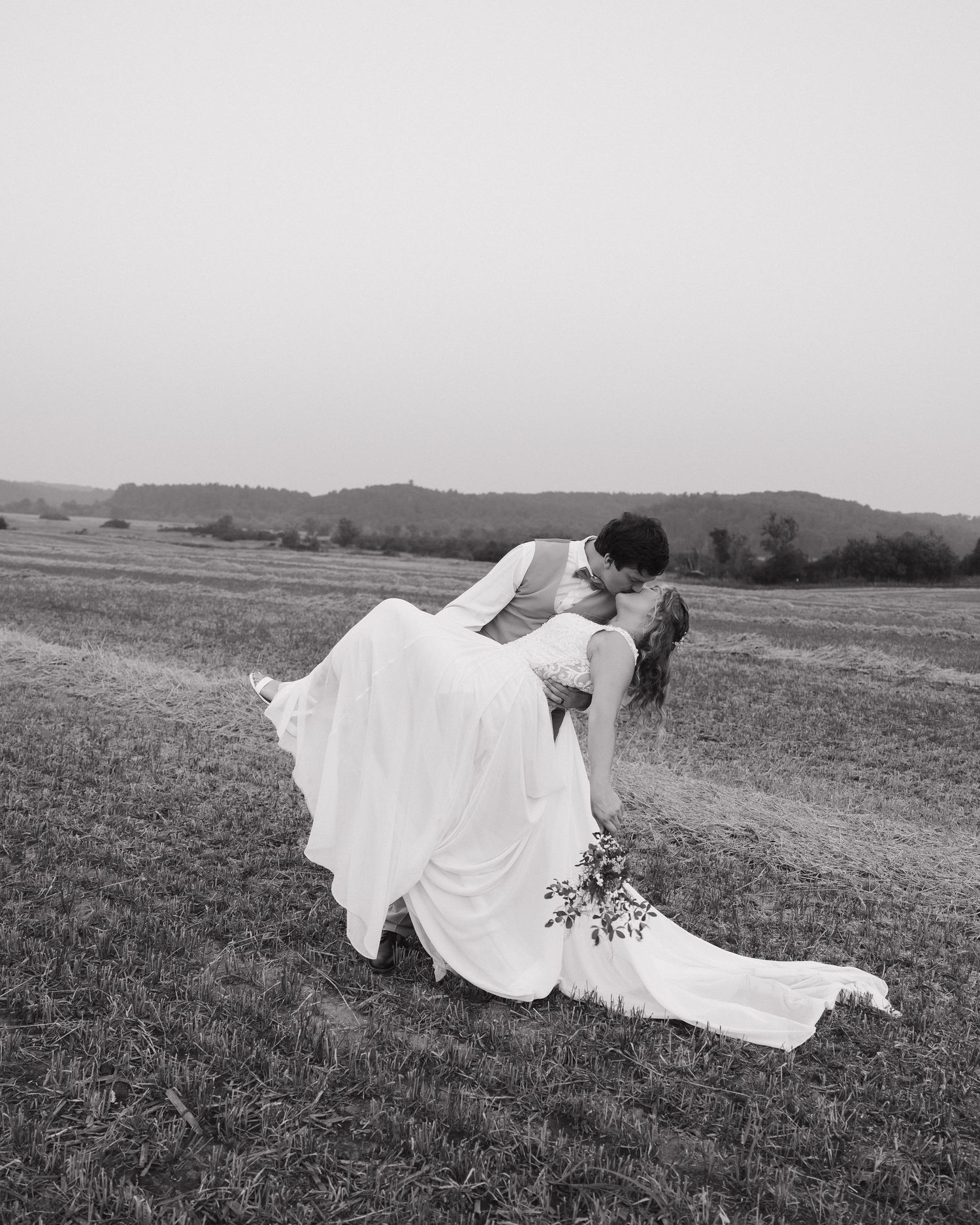 A romantic black and white photo of a newlywed couple embracing in a scenic hilltop meadow on their wedding day.