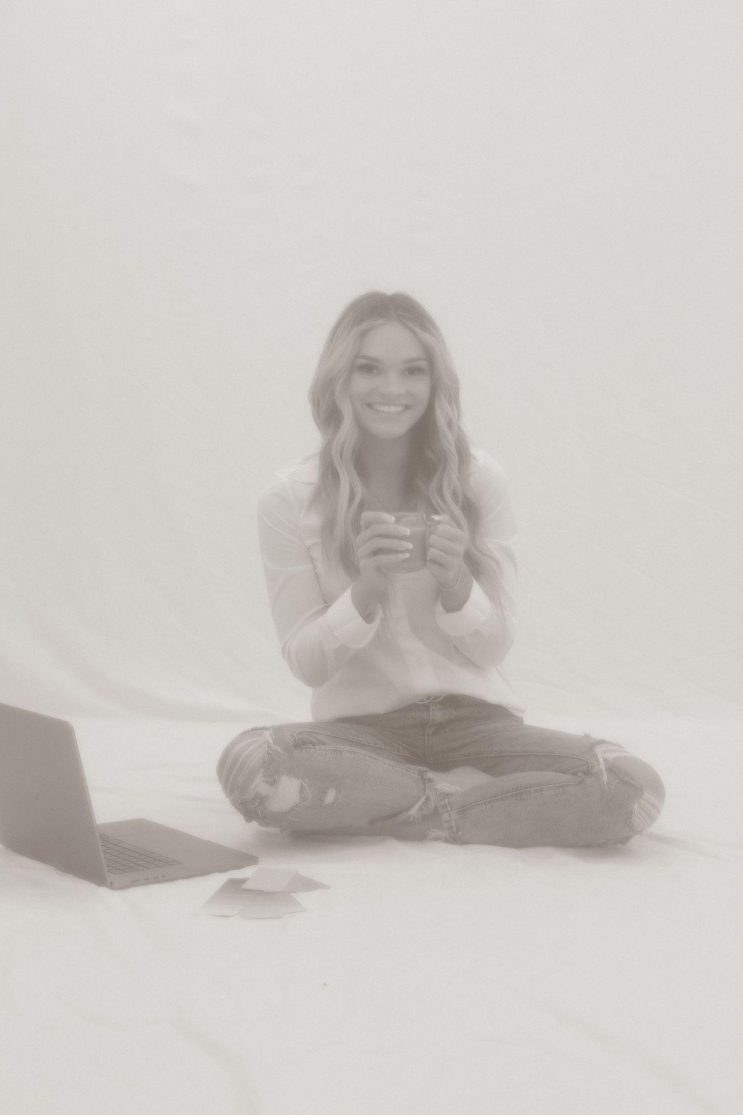 A casual portrait of someone sitting cross-legged with a laptop in a bright white studio setting.