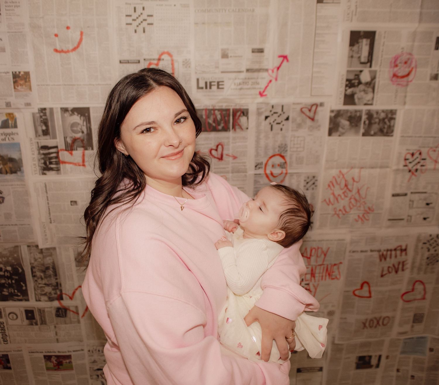 Woman in pink sweater holding baby against newspaper-covered wall with heart decorations.