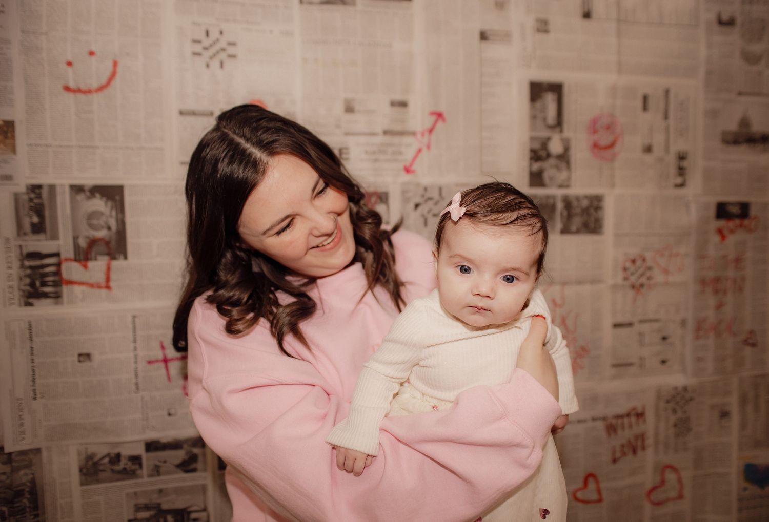 Mother in pink sweater holding baby against decorated newspaper wall background.