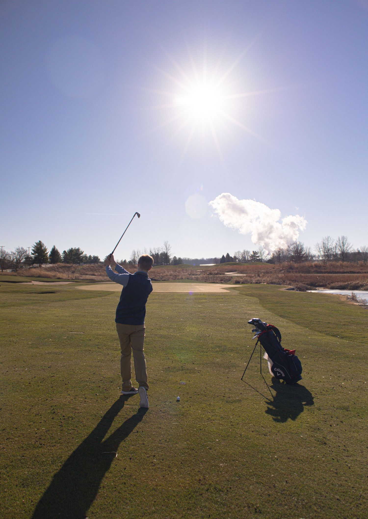 A golfer takes a swing on a sunny day with their golf bag casting a long shadow on the green.