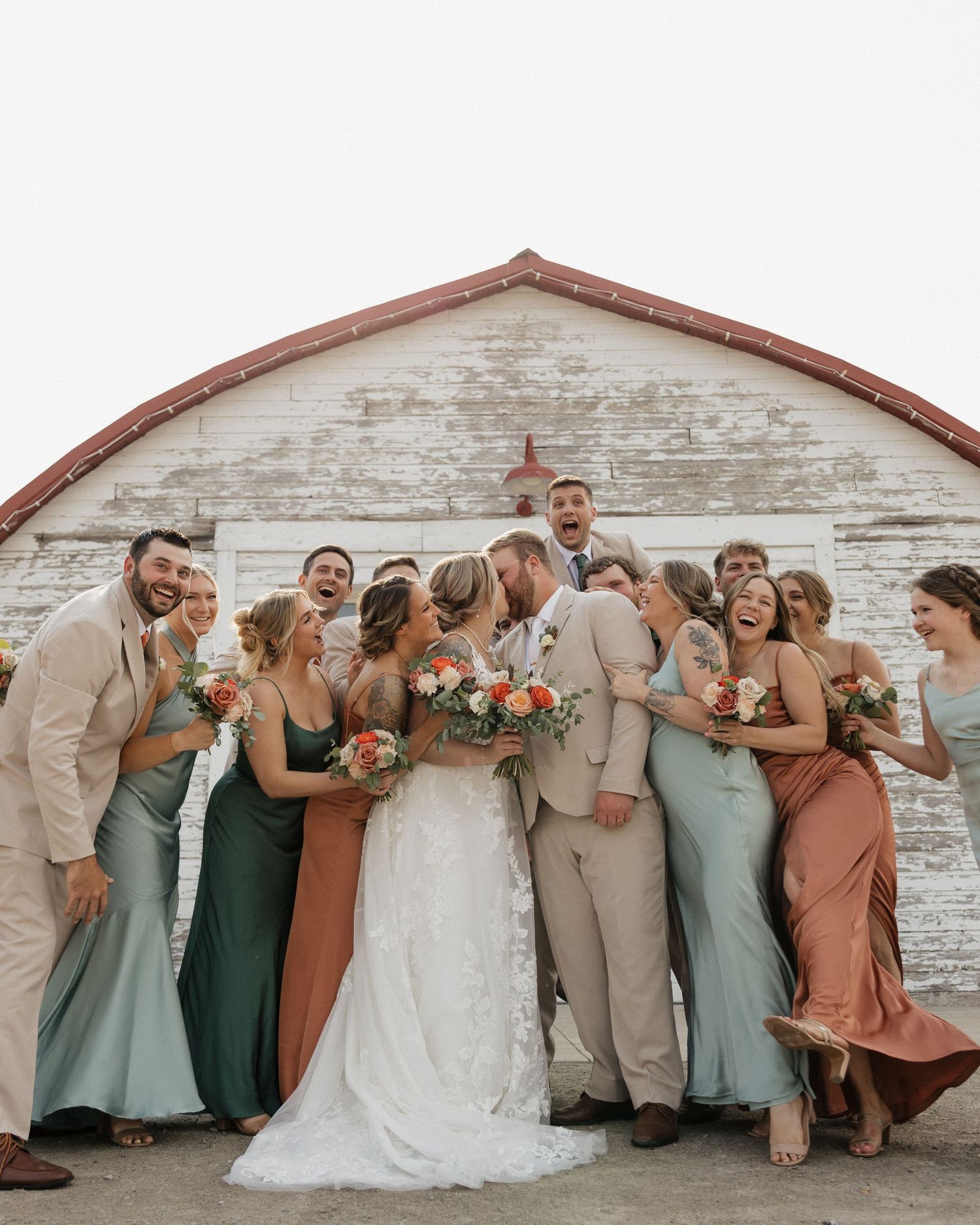 A wedding party lineup standing against a rustic white barn wall posing playfully in sage green and terracotta dresses.