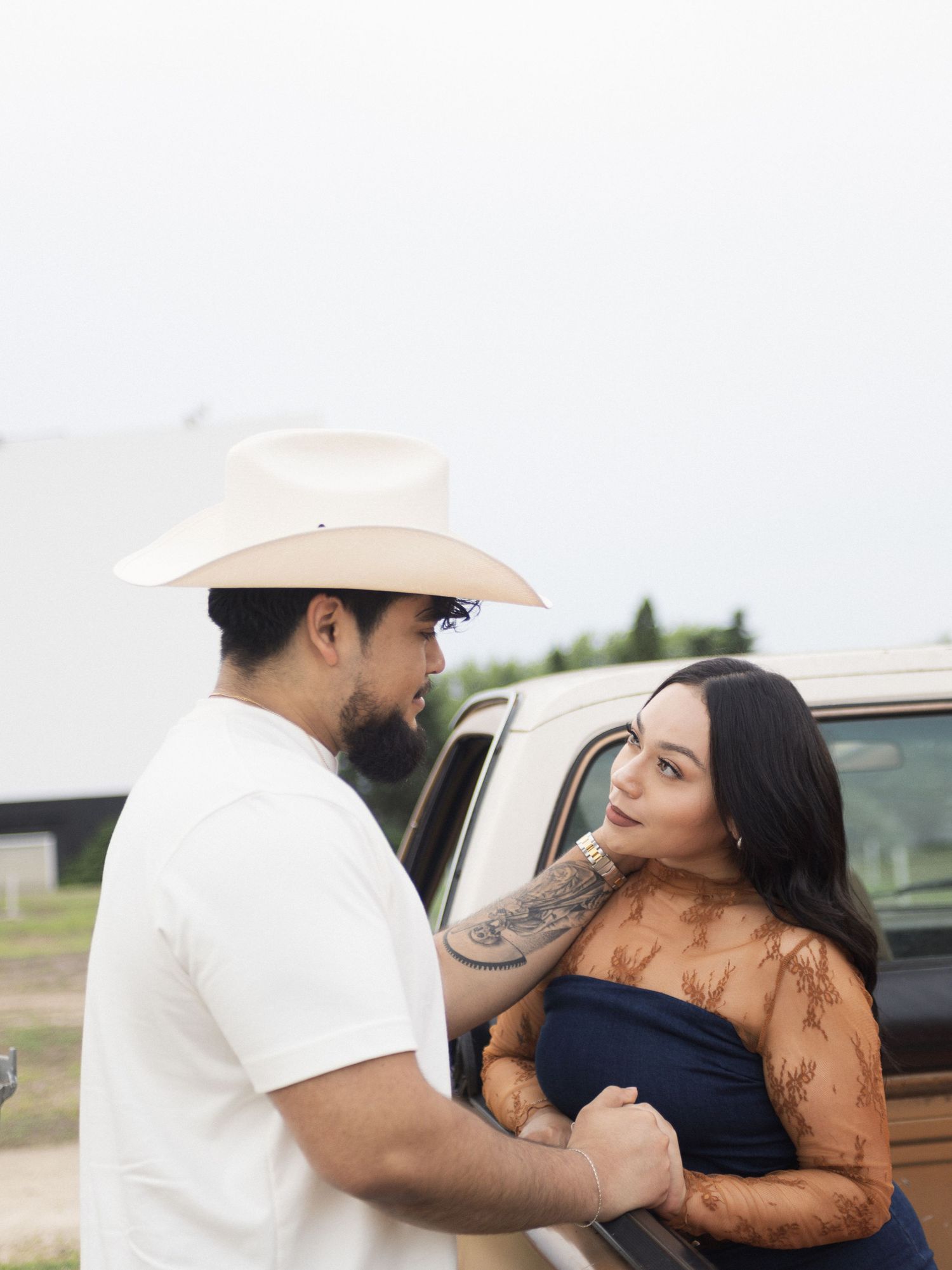 A couple in an affectionate embrace stand by a truck, with one person wearing a cowboy hat and white shirt.