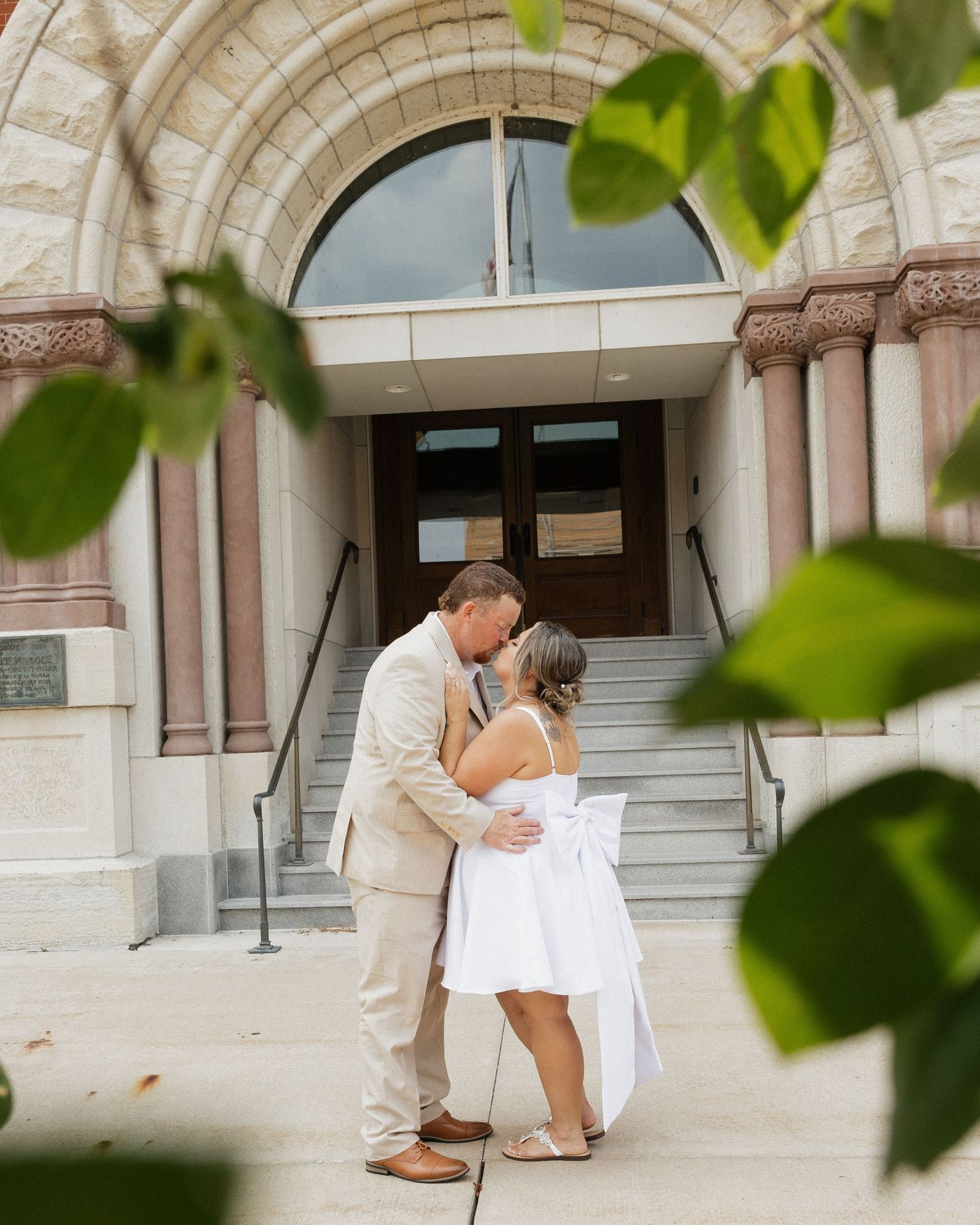 Two people share a romantic kiss on the steps of a historic stone building with an arched entryway framed by green leaves.