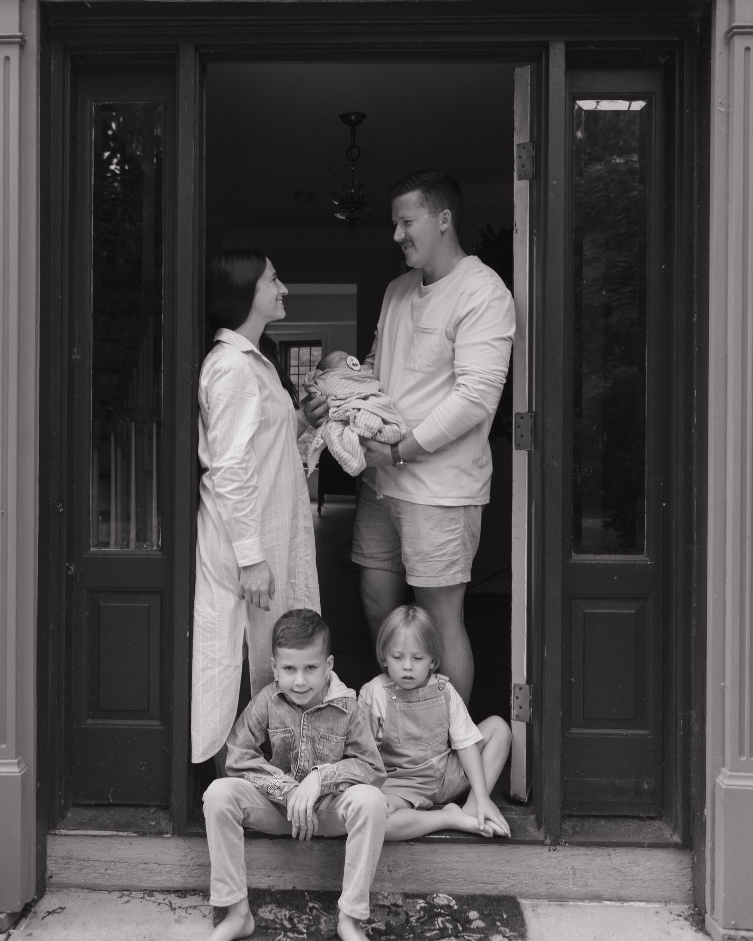 A monochrome family portrait showing two adults and two children relaxing in a doorway of their home.