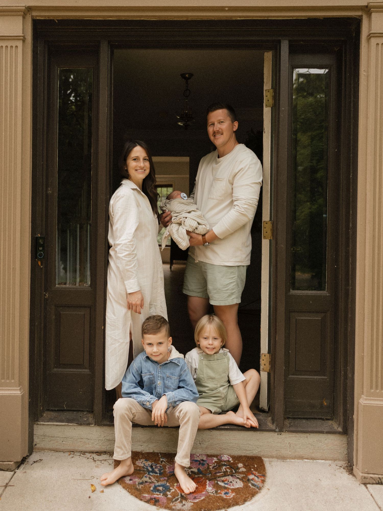 Family standing in doorway of classic home wearing coordinated casual white and neutral outfits.
