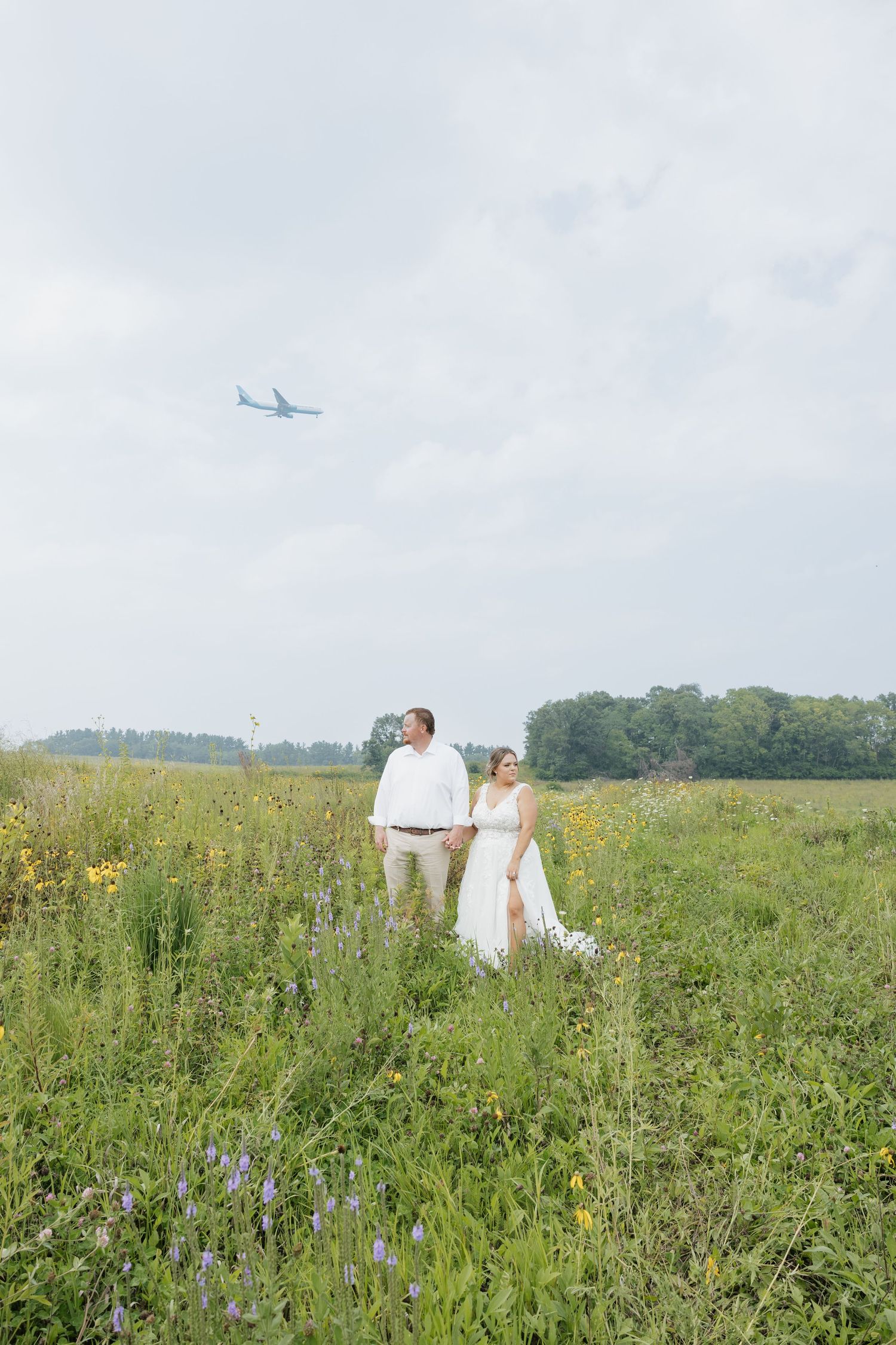 A couple walks together in a wildflower meadow as a plane flies overhead in the cloudy summer sky.