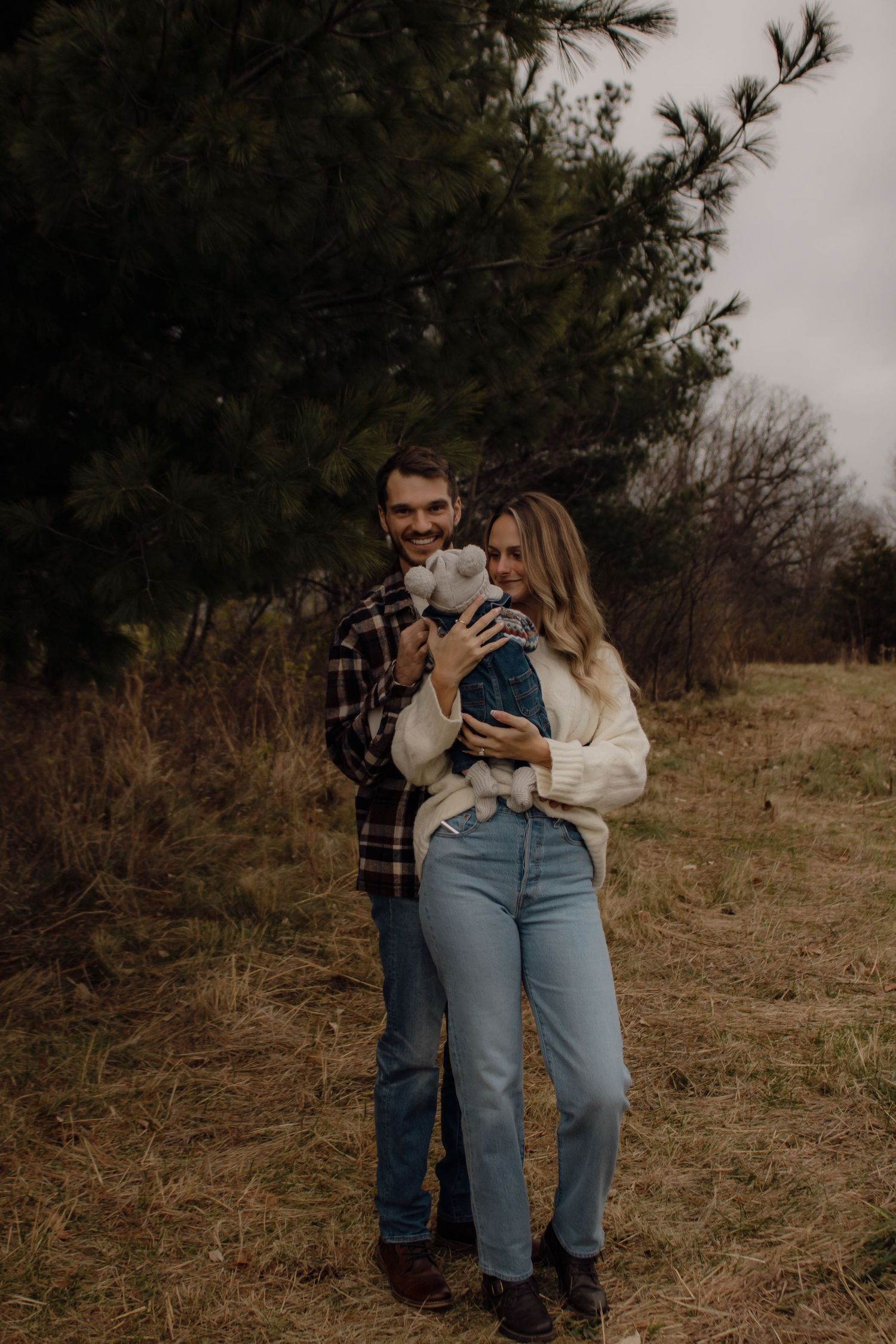 A couple in casual attire stands together outdoors holding their small dog against an earthy autumn backdrop.