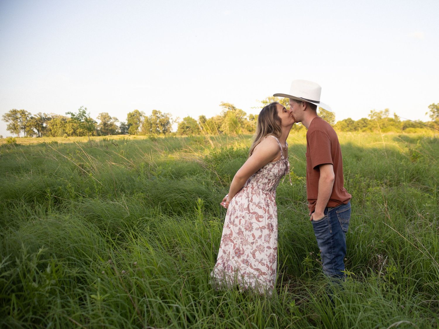 Two people share a romantic moment while standing in a tall grass field at sunset with golden sunlight.