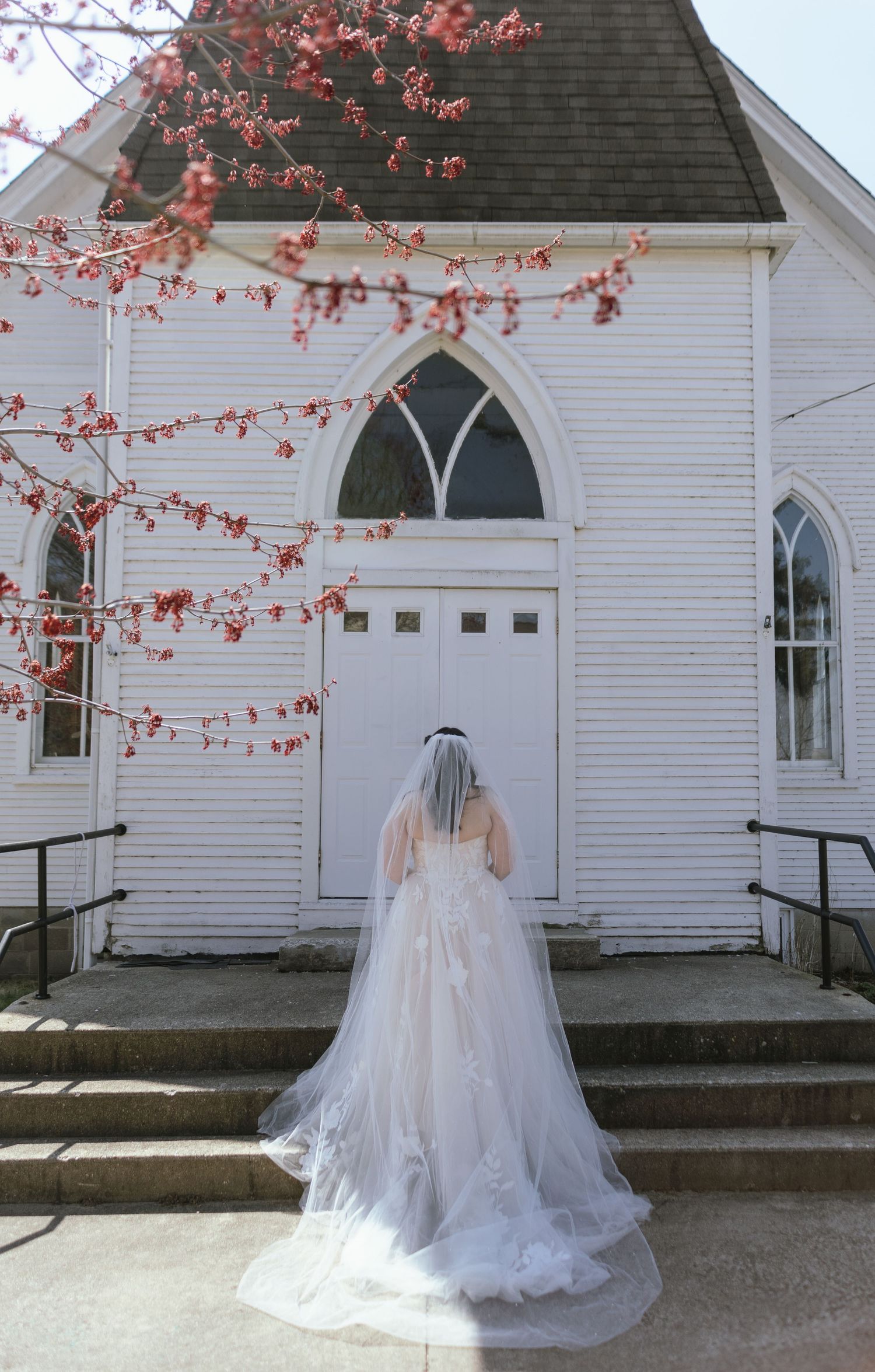 A bride in a flowing white gown and veil standing before a white church with blooming spring branches.