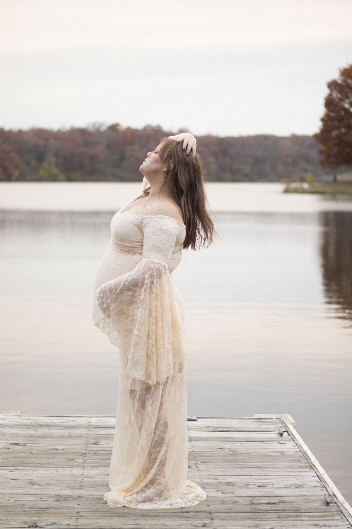 Figure in lace dress stands on wooden dock overlooking serene lake at sunset.