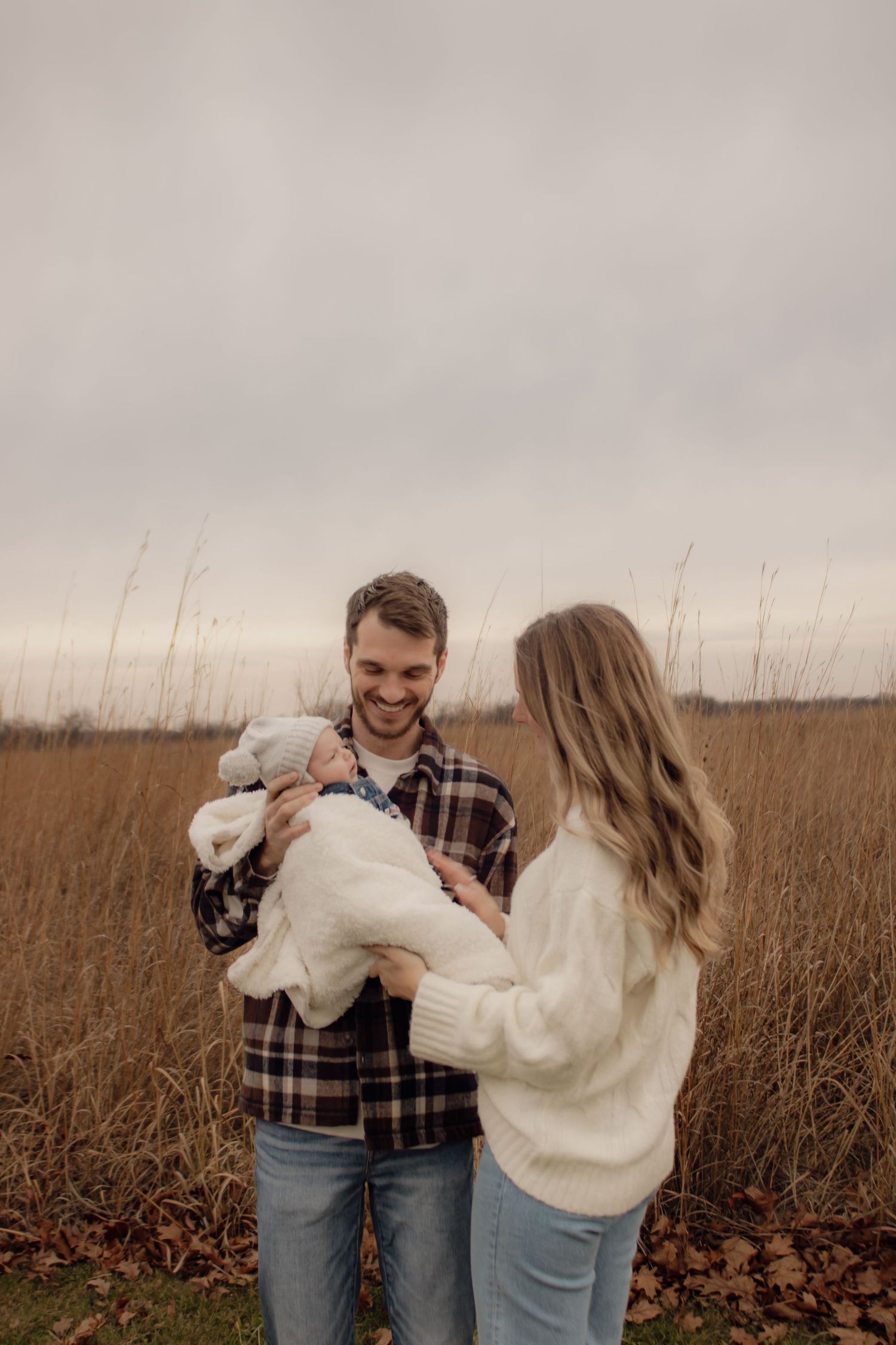 A couple holding a small white dog stands together in a field of tall brown grass on an overcast day.