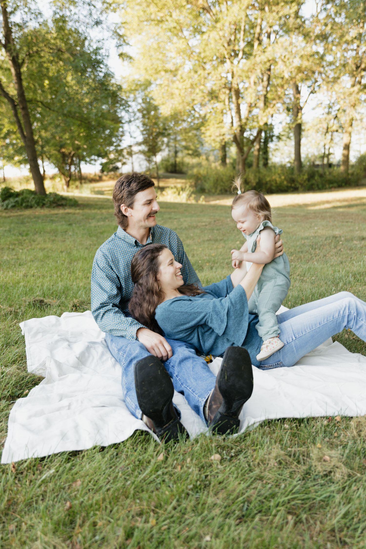 A couple relaxes and laughs together while lying on a white blanket in a sunny grassy field on an autumn day.