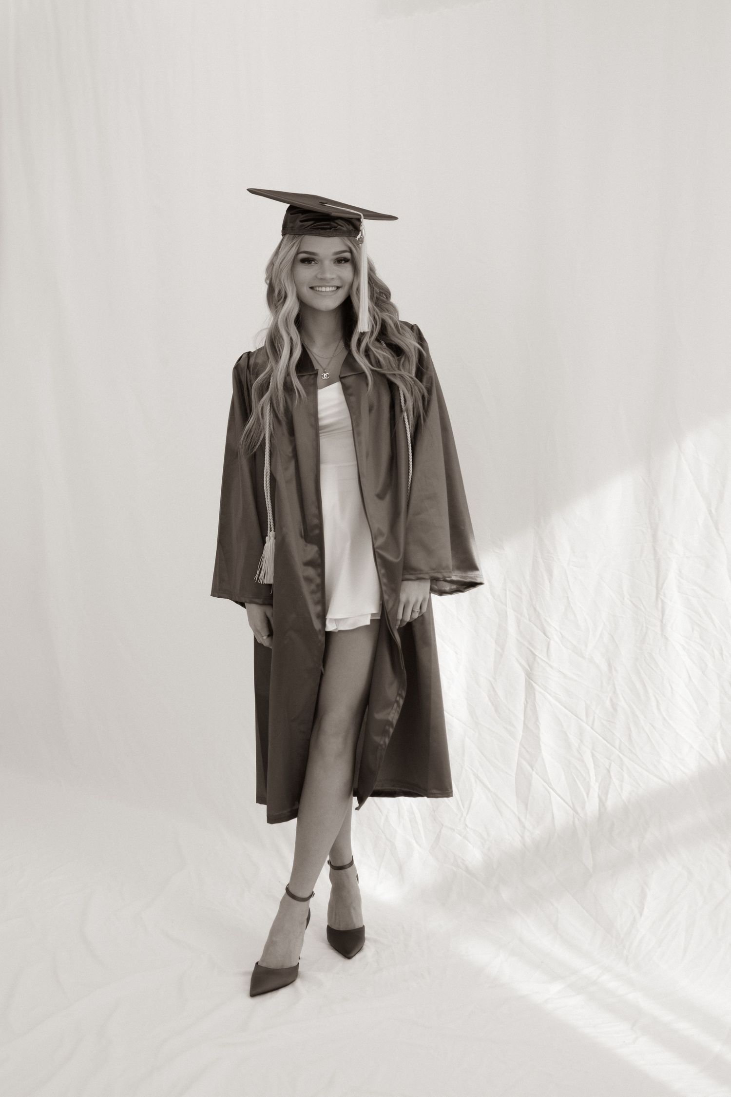 A graduate poses in cap and gown against a white backdrop in a classic black and white portrait.