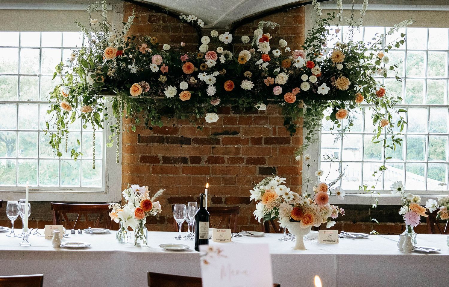 Elegant wedding reception table with hanging floral arrangement of peach and white blooms above brick wall.