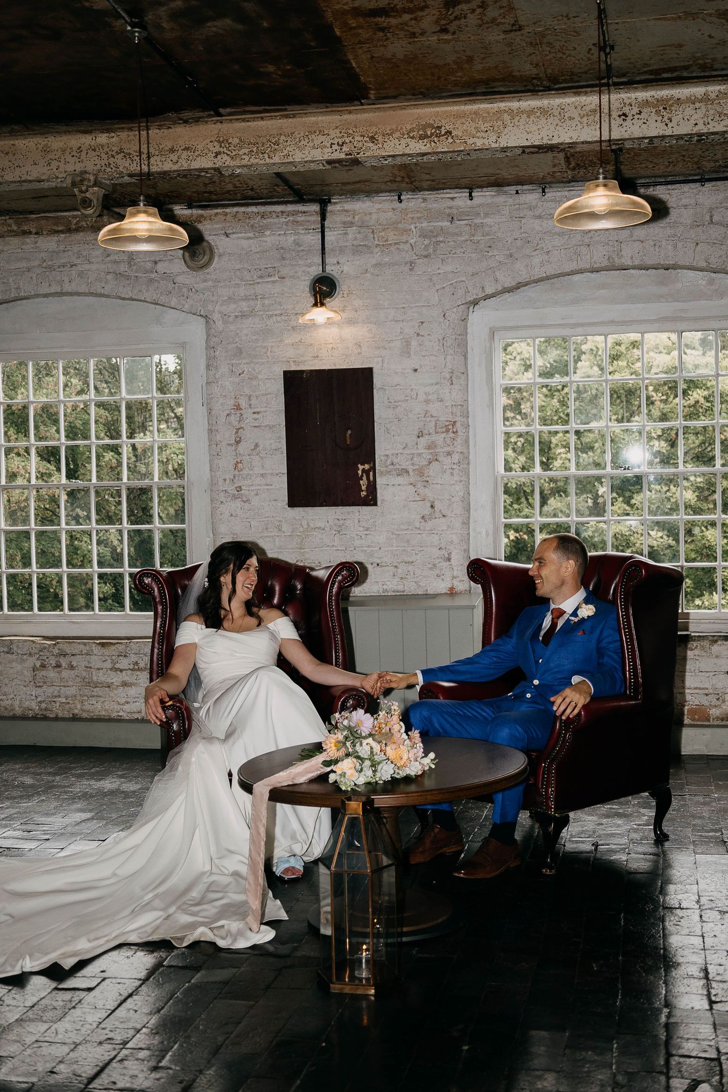 Bride and groom sitting on vintage leather chairs holding hands at an industrial wedding venue with white brick walls.