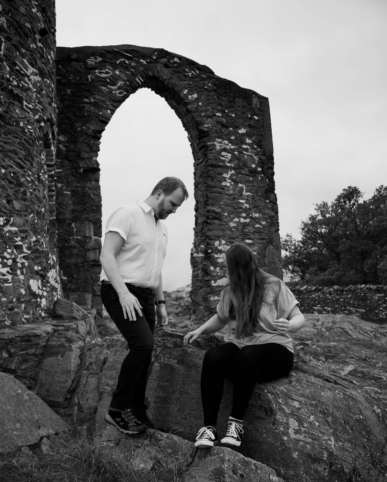 Black and white photo of a couple at ancient stone ruins with an arched doorway, woman sitting on rocks while man stands nearby.