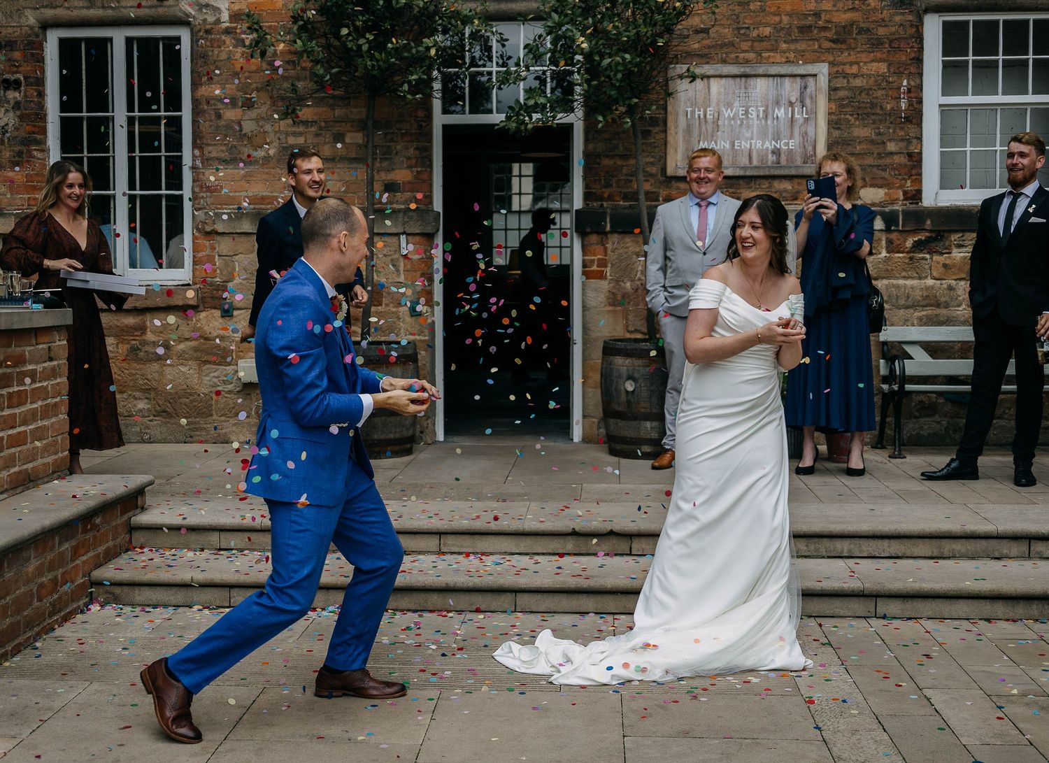 Bride and groom celebrate with colorful confetti outside a brick venue, guests watching joyfully during outdoor wedding ceremony.