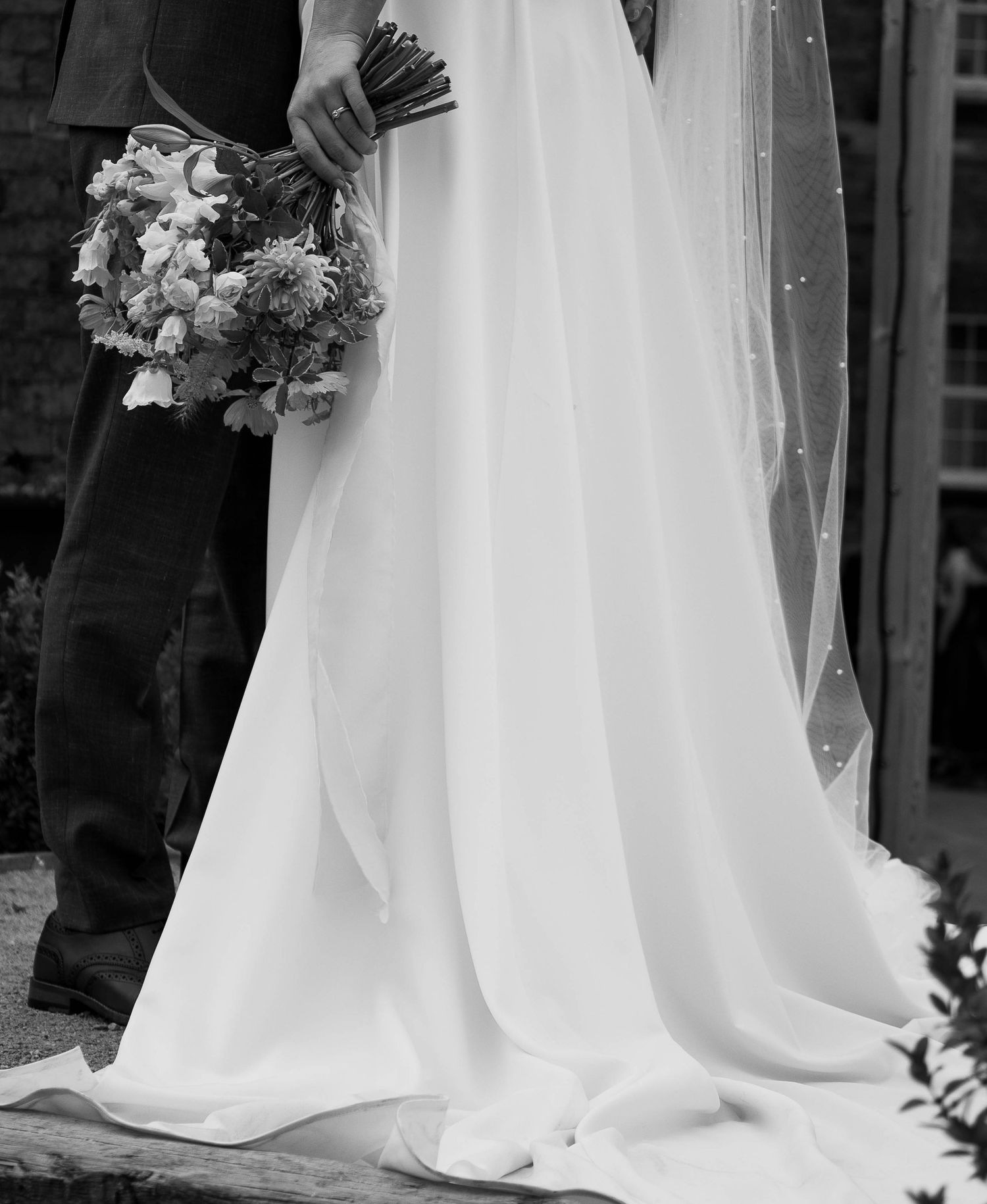 Black and white photo of a bride in a flowing gown holding a bouquet, standing beside a groom in a suit.