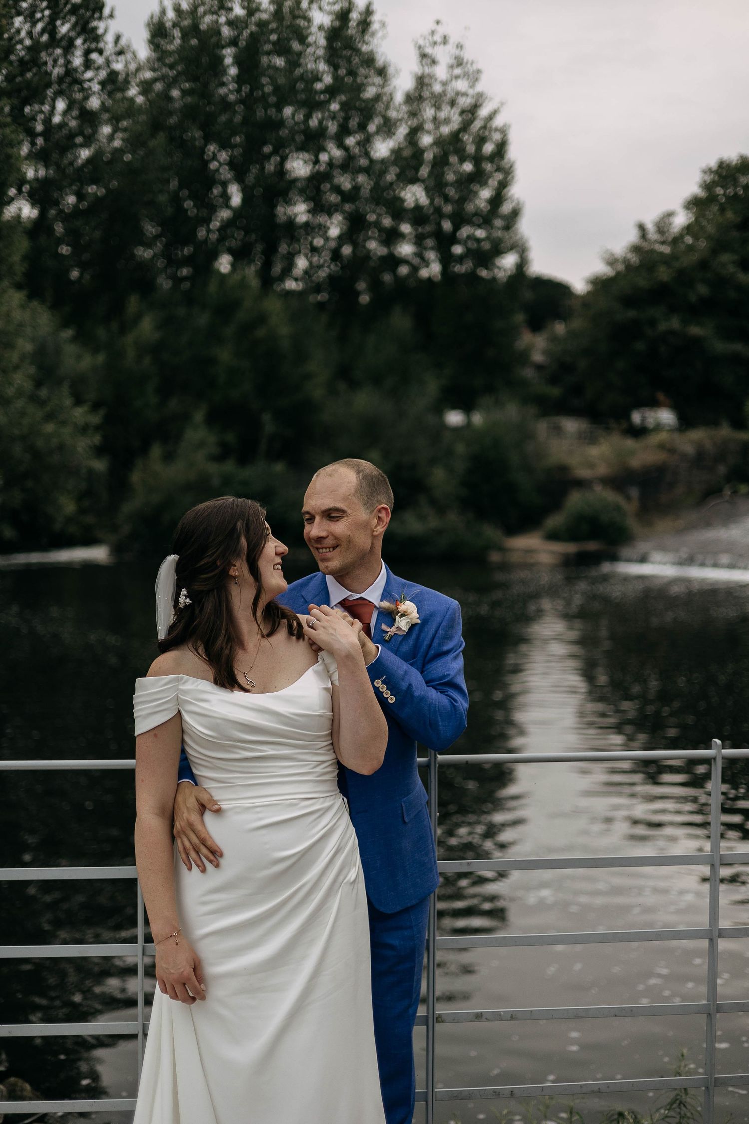 Bride in white off-shoulder gown and groom in blue suit share a romantic moment on a bridge by a river.