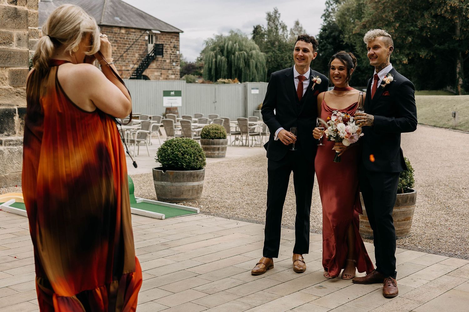 Wedding photographer capturing a bride and groom posing with a guest outdoors at a rustic stone venue.